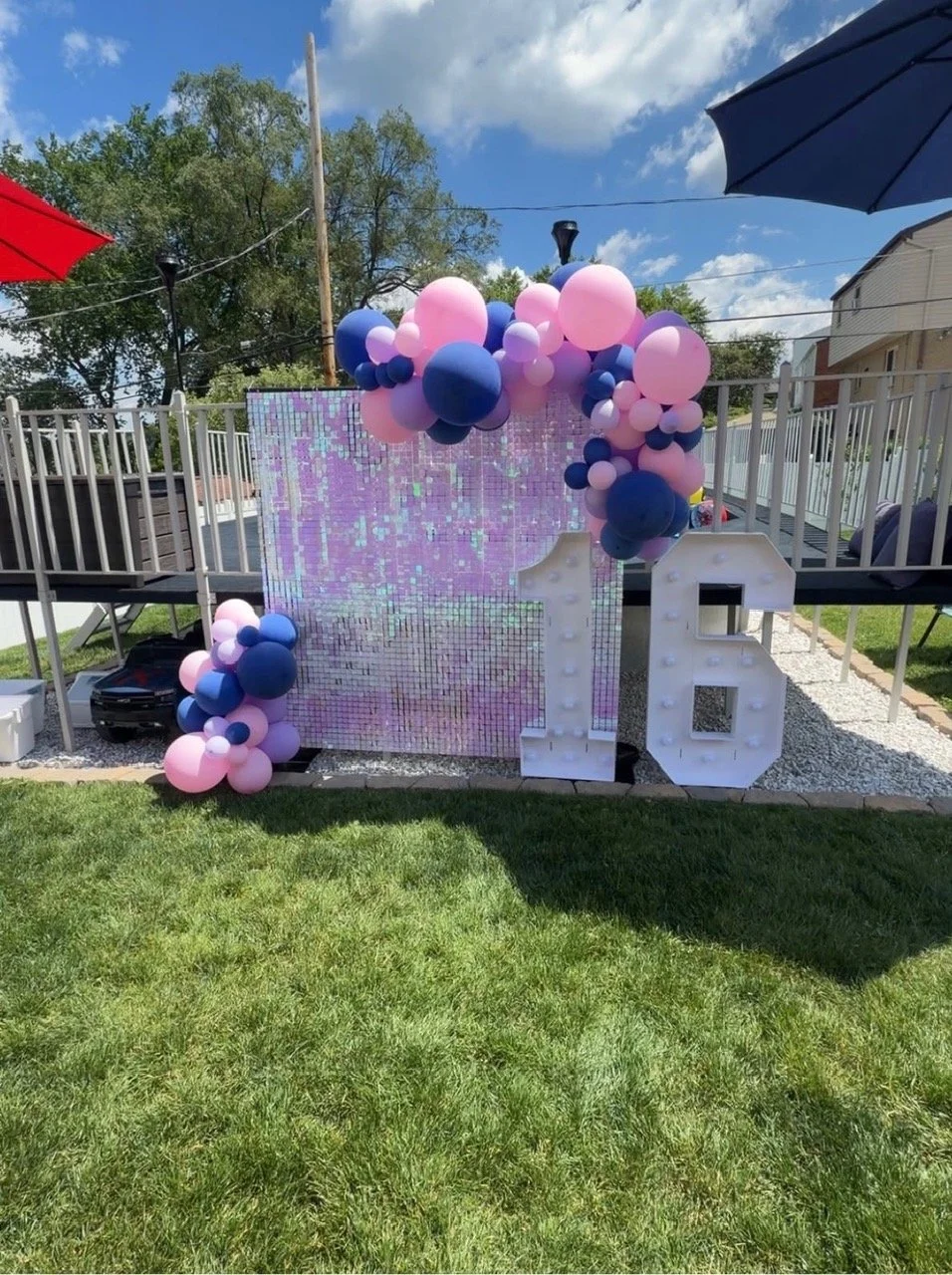 Decorative birthday celebration setup with large number 16, pink and purple balloons, a mirrored backdrop, and umbrellas providing shade in a backyard.