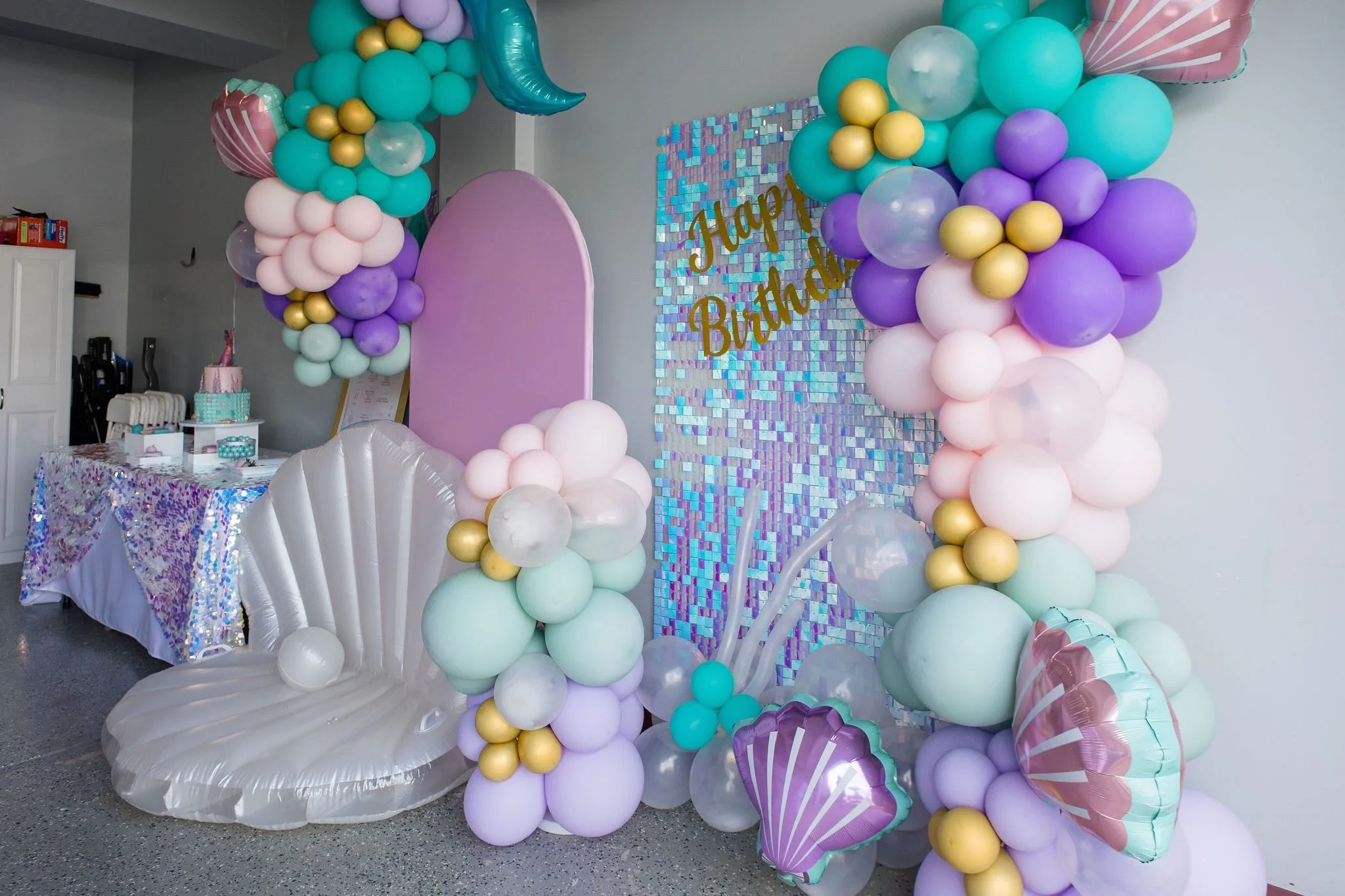 Colorful balloon arch and decorations for a birthday celebration, including pastel and metallic balloons, a shimmering tablecloth, and a sign that says 'Happy Birthday'.