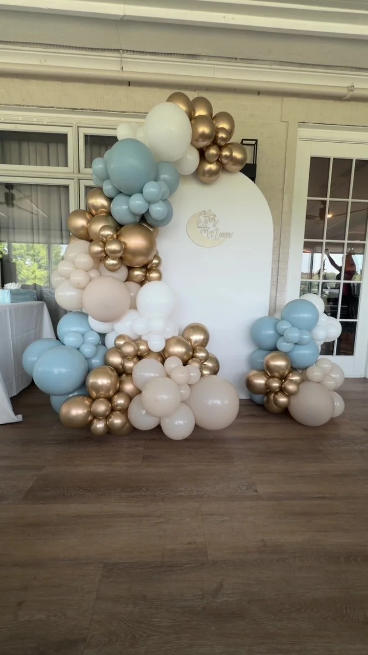 Balloon arch and clusters in pastel blue, white, and gold in front of a white backdrop with a moon and unicorn design for a celebration or event.