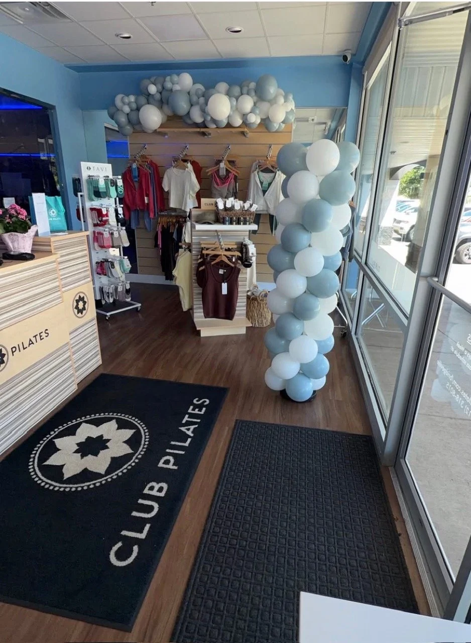 Interior of a fitness or wellness store with Balloon decorations, clothing on display, and a Club Pilates branded mat at the entrance.