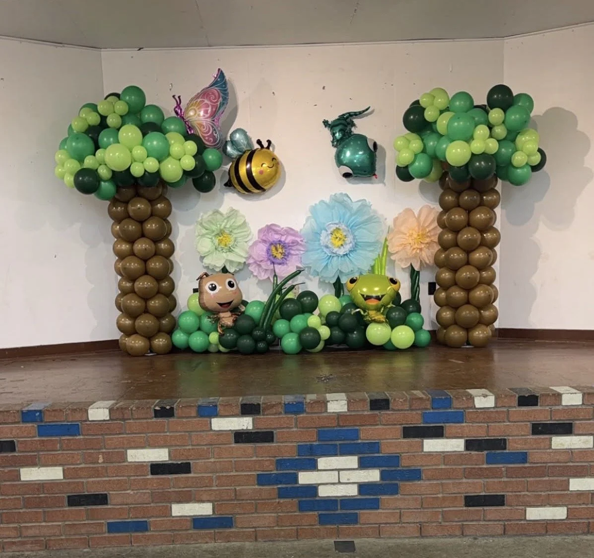 Decorative display with balloon trees, insects, and flowers on a stage, with brick wall in front.
