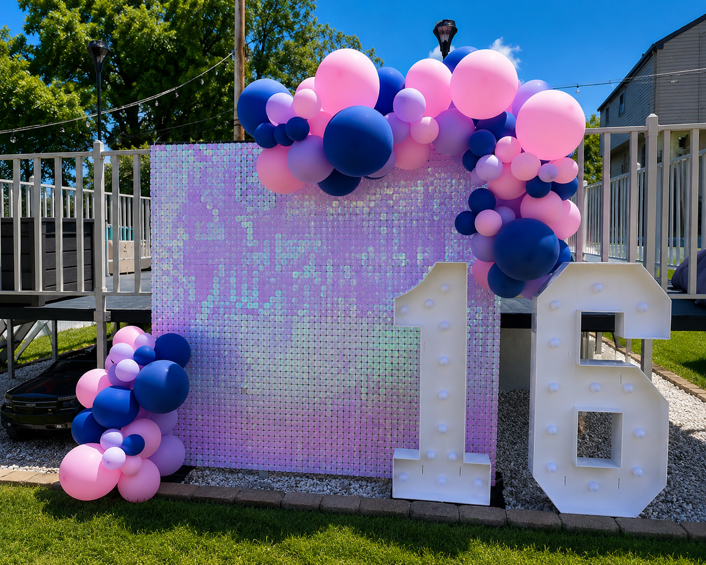 Decorative outdoor birthday backdrop with pink, purple, and blue balloons, a large white number 18, and an iridescent square panel on a grassy area under a blue sky.