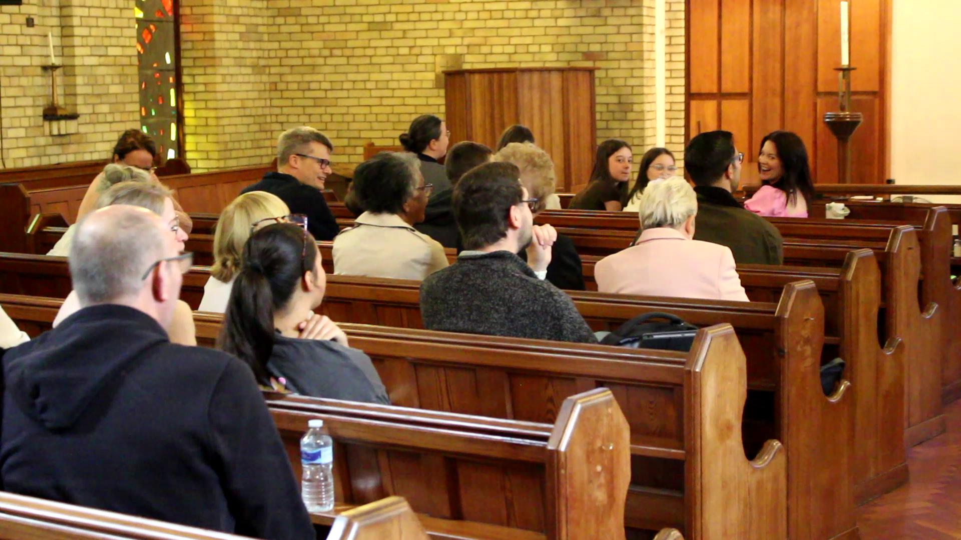 People seated in wooden pews inside a church, engaged in conversation and listening to a speaker.