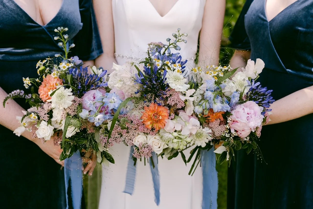 Close-up of a wedding bouquet featuring pink, white, purple, and blue flowers with greenery, held by women in formal dresses, possibly at a wedding or special event.