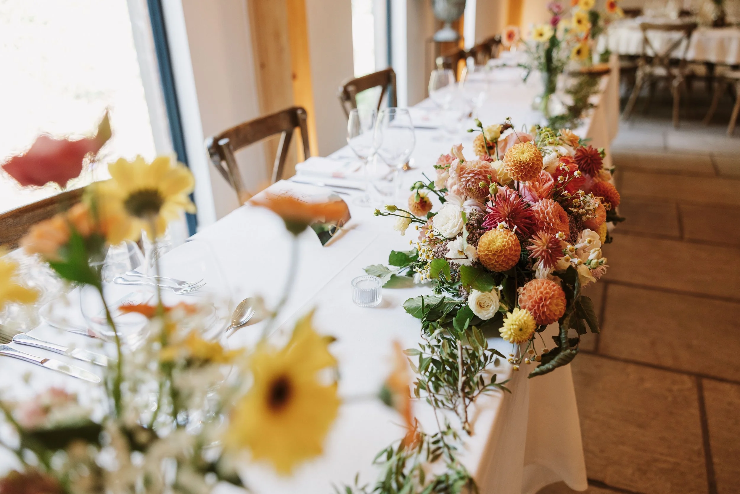 A close-up of a floral centerpiece on a banquet table, with a larger floral arrangement in the background, set in a well-lit room with wooden elements and large windows.