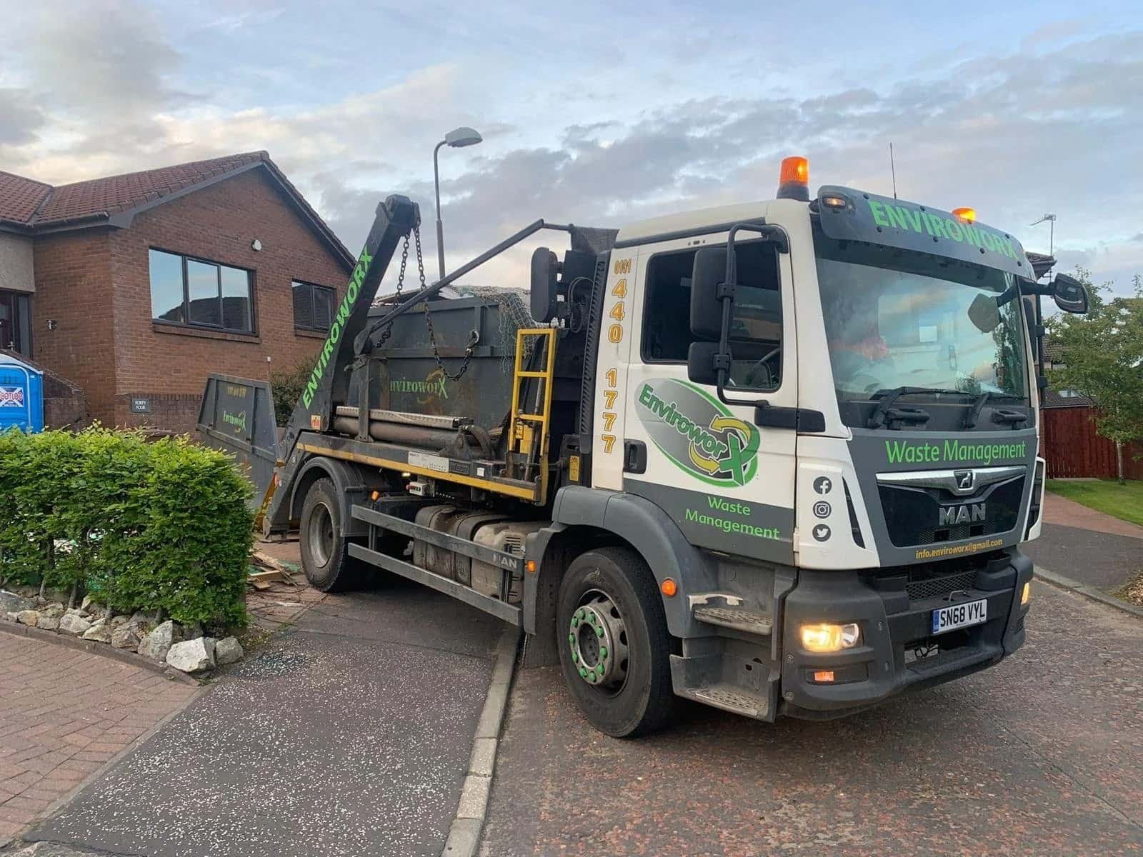 White MAN truck with green ‘Enviroworx Waste Management’ branding and phone number 040 1777, parked on a residential street with a hydraulic arm and skip container.