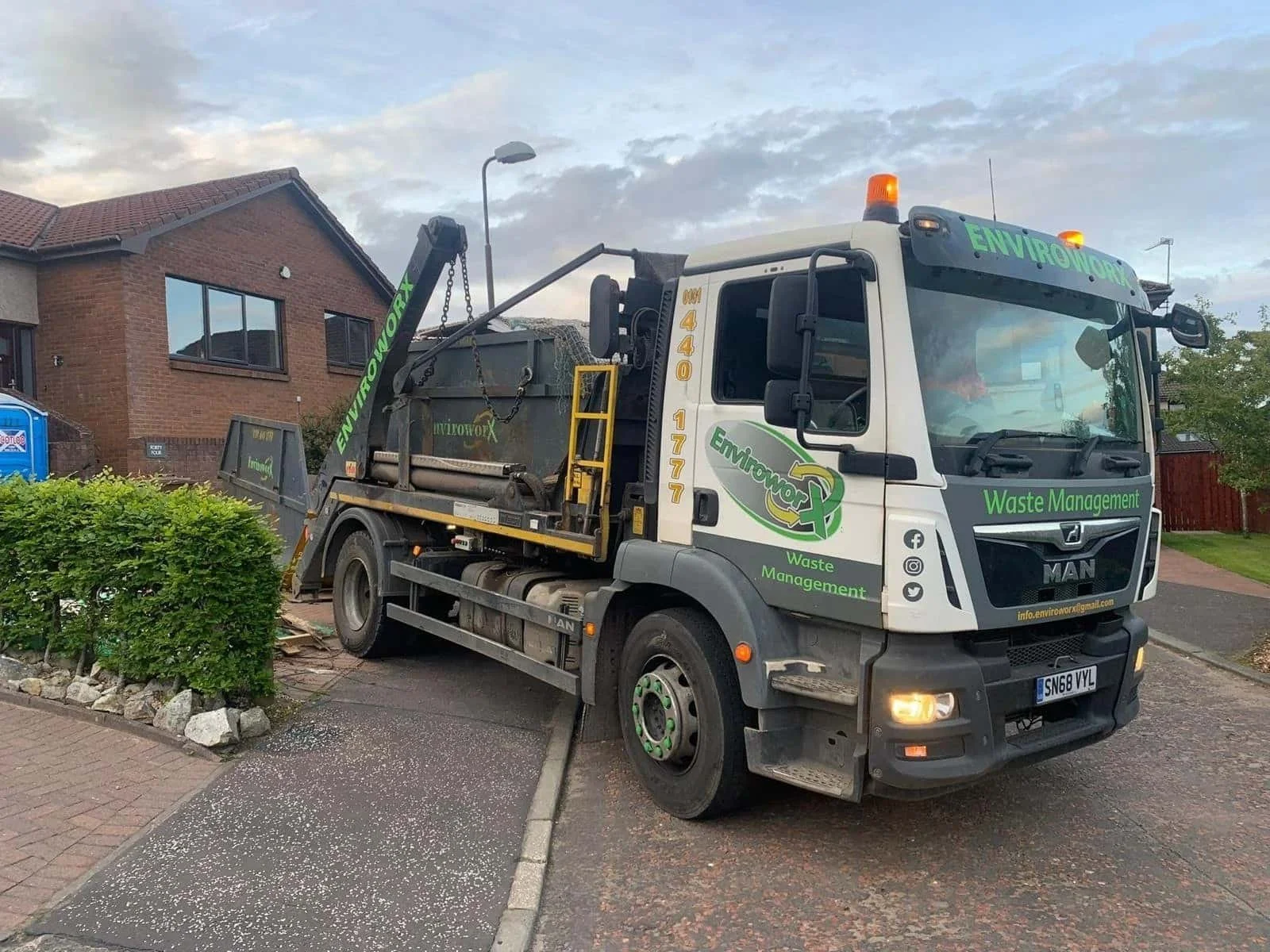 White MAN truck with green ‘Envirowork Waste Management’ branding and phone number 040 1777, parked on a residential street with a hydraulic arm and skip container.
