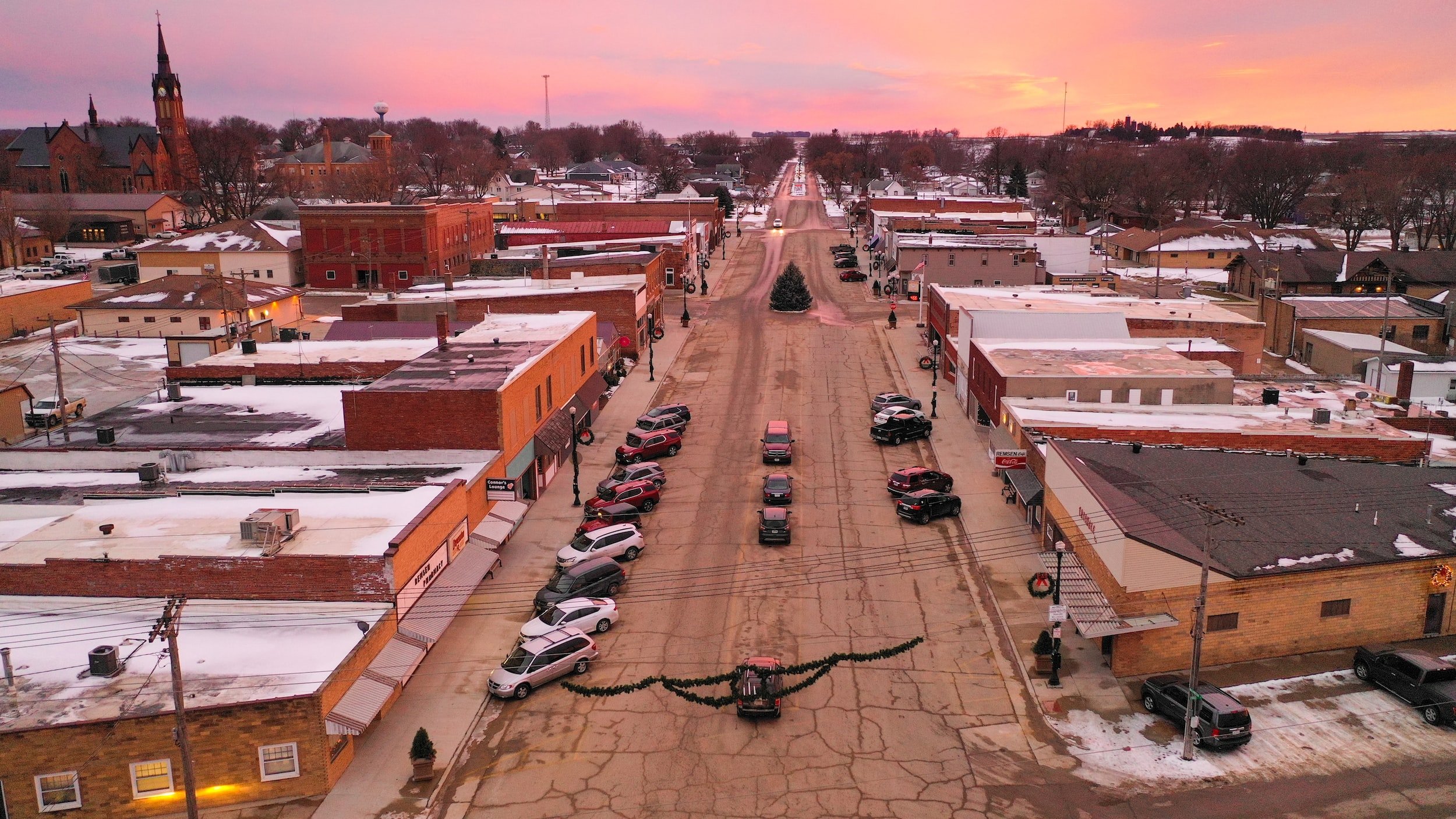 Town image - a broken asphalt wide street with angled parking, low brick buildings lining the sides.