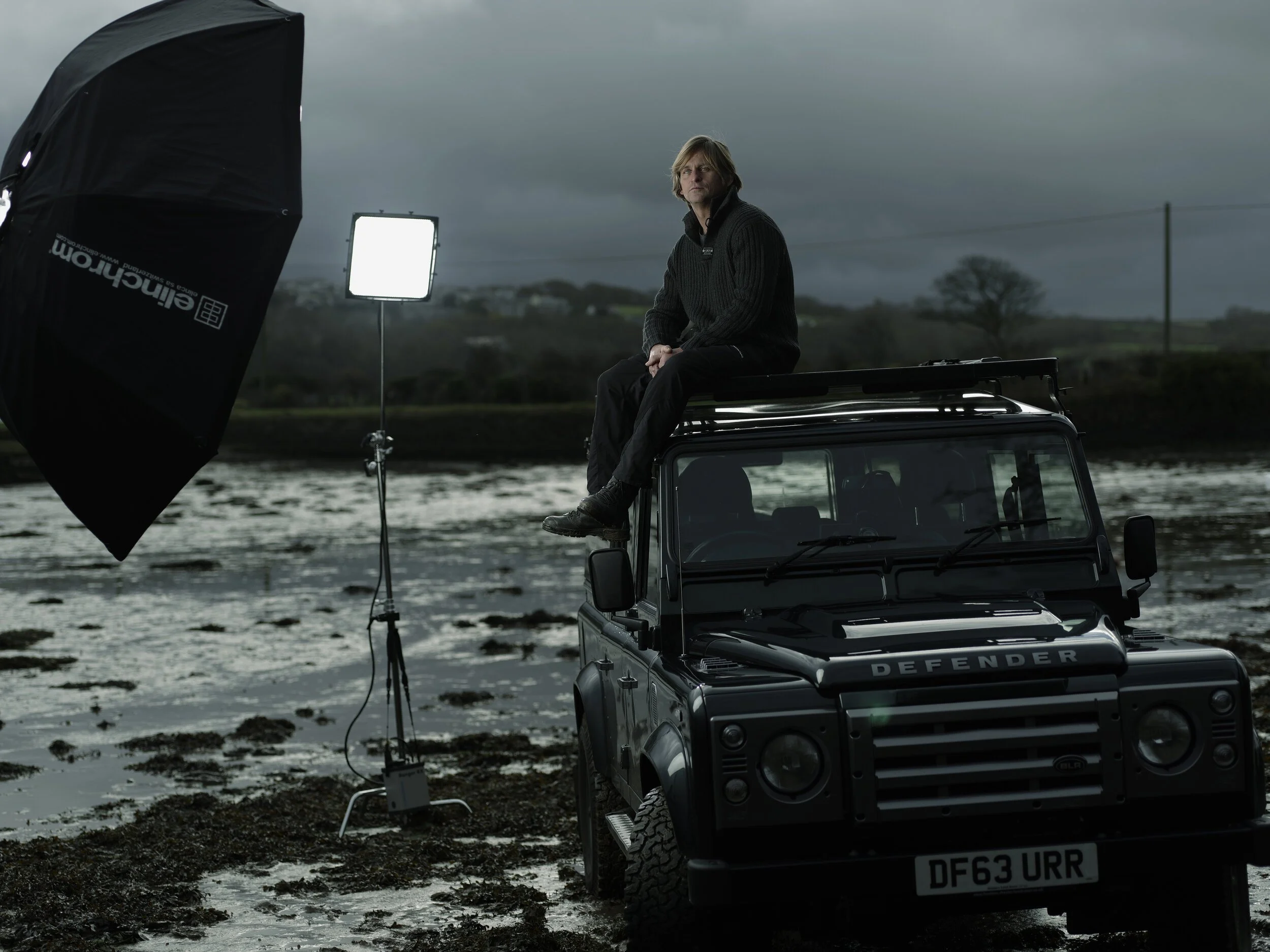 A man sitting on top of a Land Rover vehicle in an outdoor setting with a cloudy sky, near water and mud, during overcast weather. Photography equipment and lighting setup are visible.