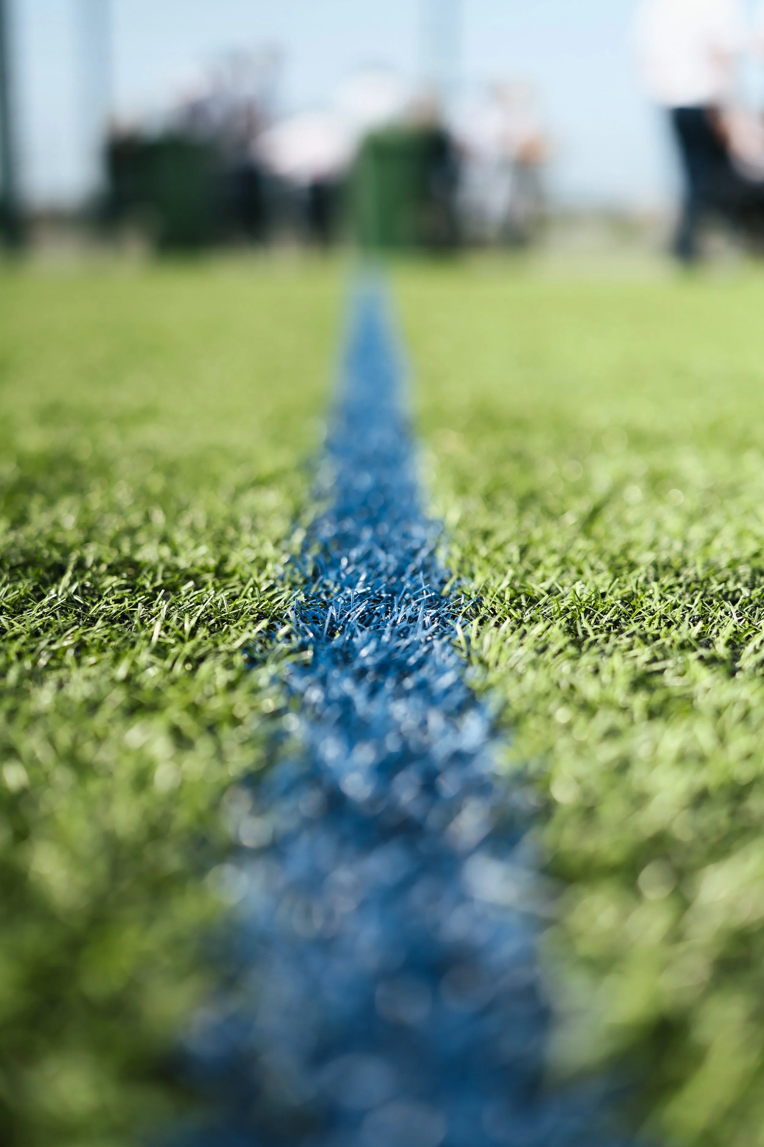 Blurred photo of a football field with a blue sideline or logo in focus, with background showing stadium seating and people.