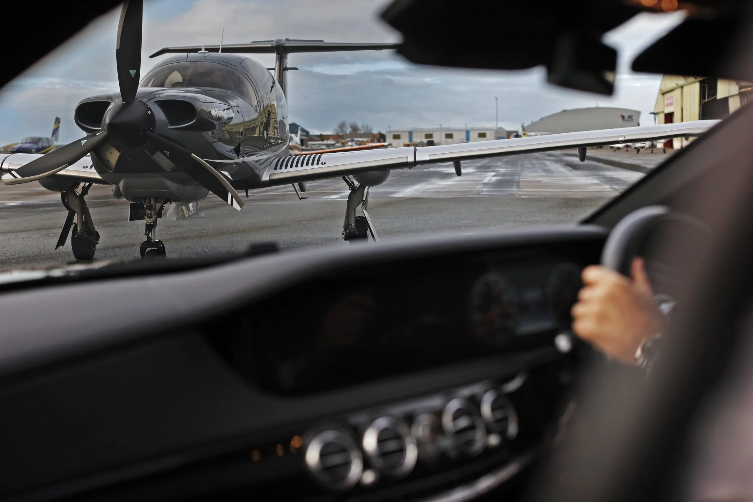 View through a car windshield showing a black aircraft parked on the tarmac, with other aircraft and airport buildings in the background.