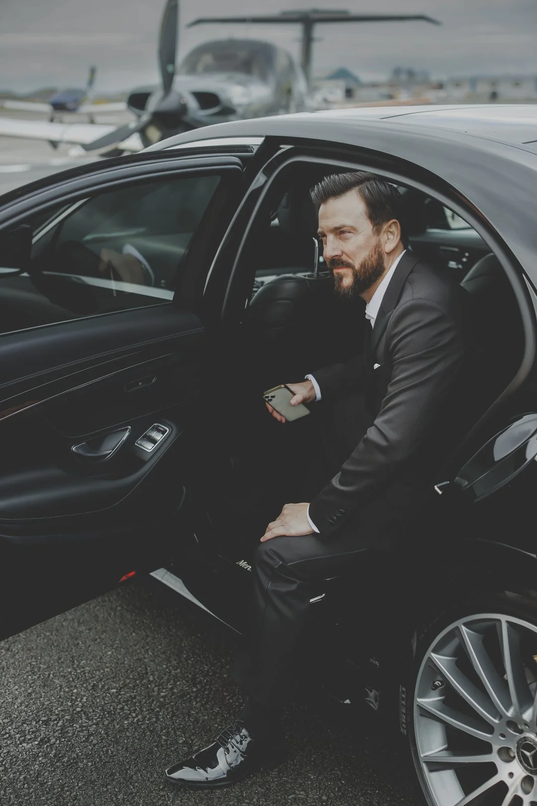Man in a suit exiting a black luxury car on an airport tarmac, with a private jet in the background.