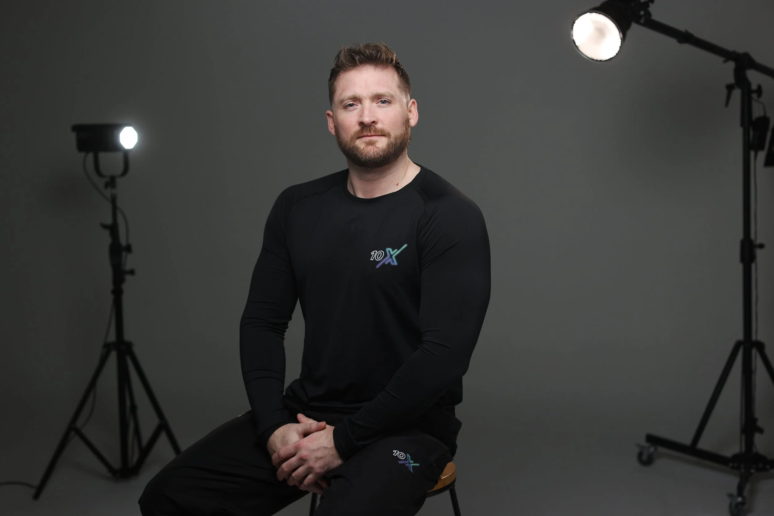 A man with a beard sitting on a wooden stool in a photography studio with professional lighting. He is wearing a black athletic shirt and black pants with a logo that says '10X'. The studio has a plain gray background and lighting equipment on tripod