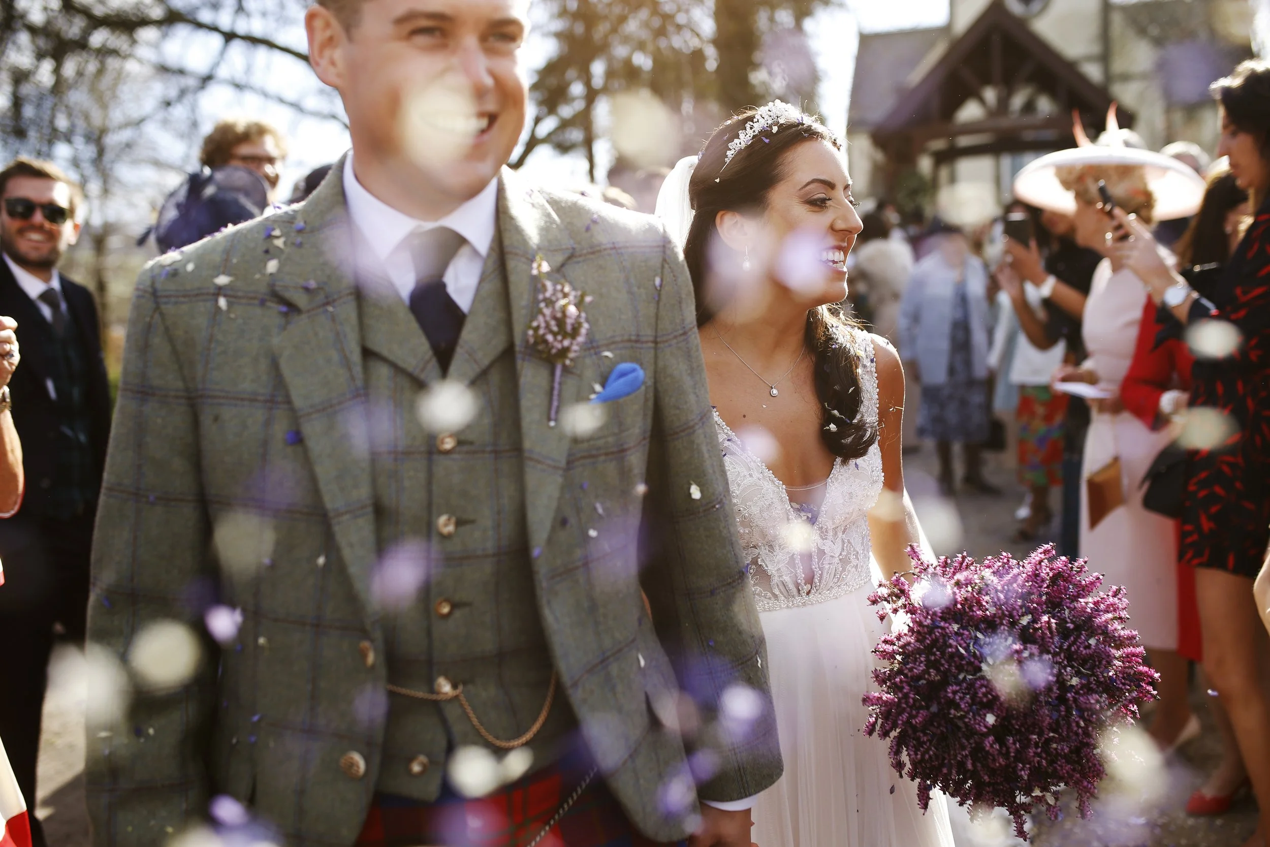 Bride and groom smiling while walking surrounded by guests, with confetti in the air and a bouquet of purple flowers.