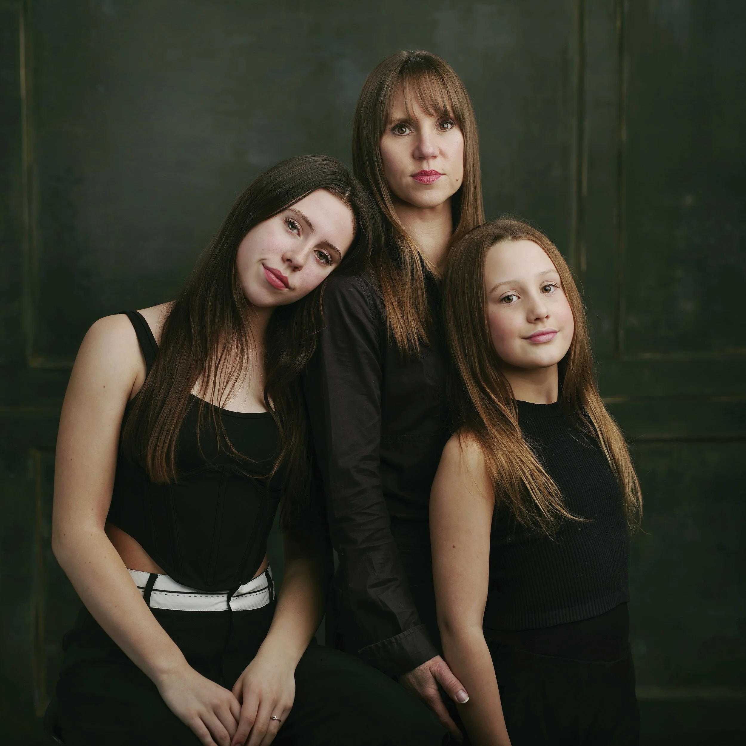 Three people posing together in front of a dark background, with similar long, straight hair, wearing black outfits. The person in the center stands while the two on either side lean slightly, creating a sense of closeness.