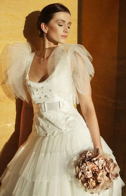 A woman in an elegant white wedding dress with tulle sleeves is standing in sunlight. She is holding a bouquet of pink flowers and looking serene and graceful.