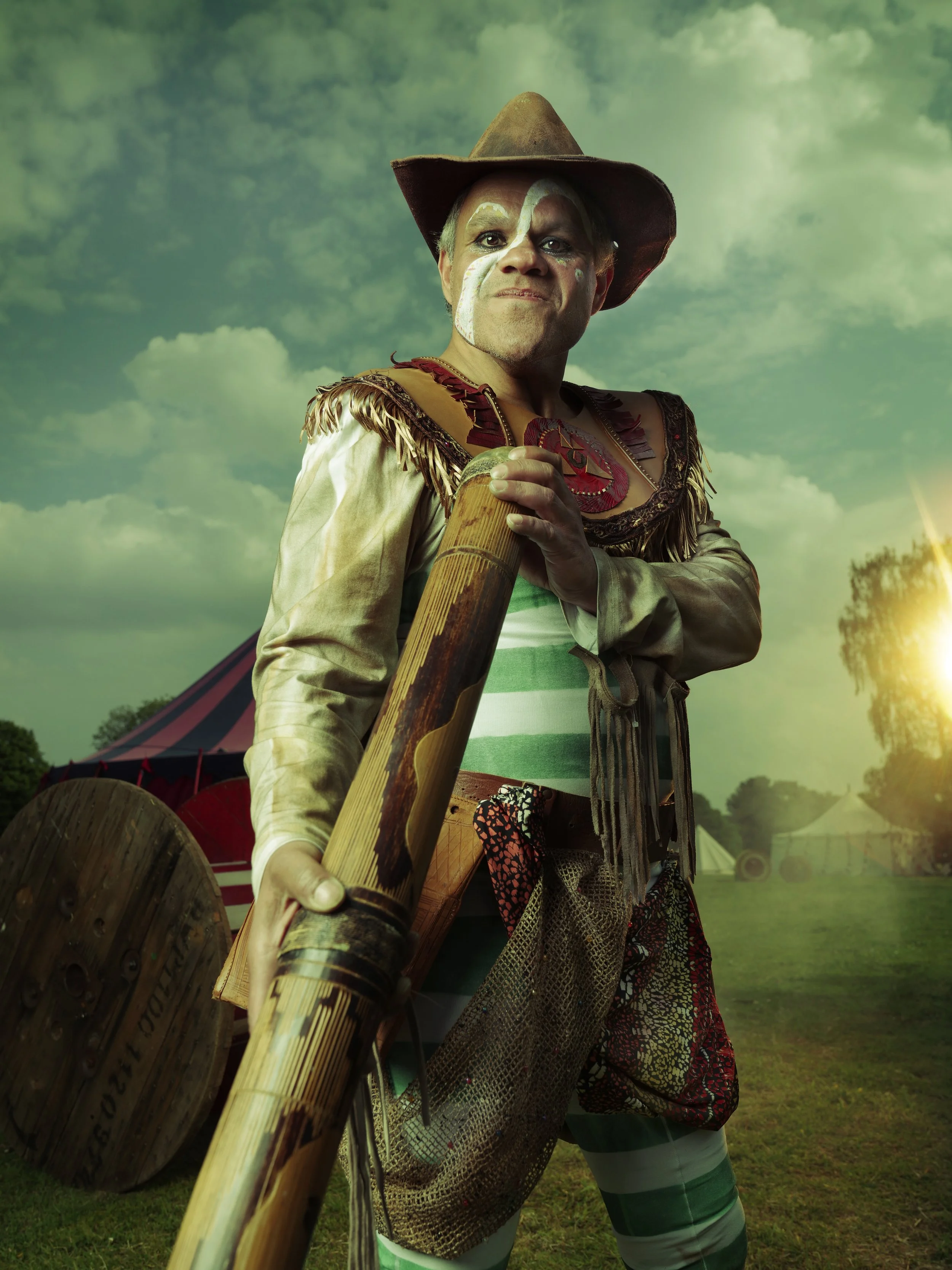 A performer in colorful costume, face paint, and a cowboy hat holding a large wooden xylophone in an outdoor festival setting with a tent and green trees in the background.