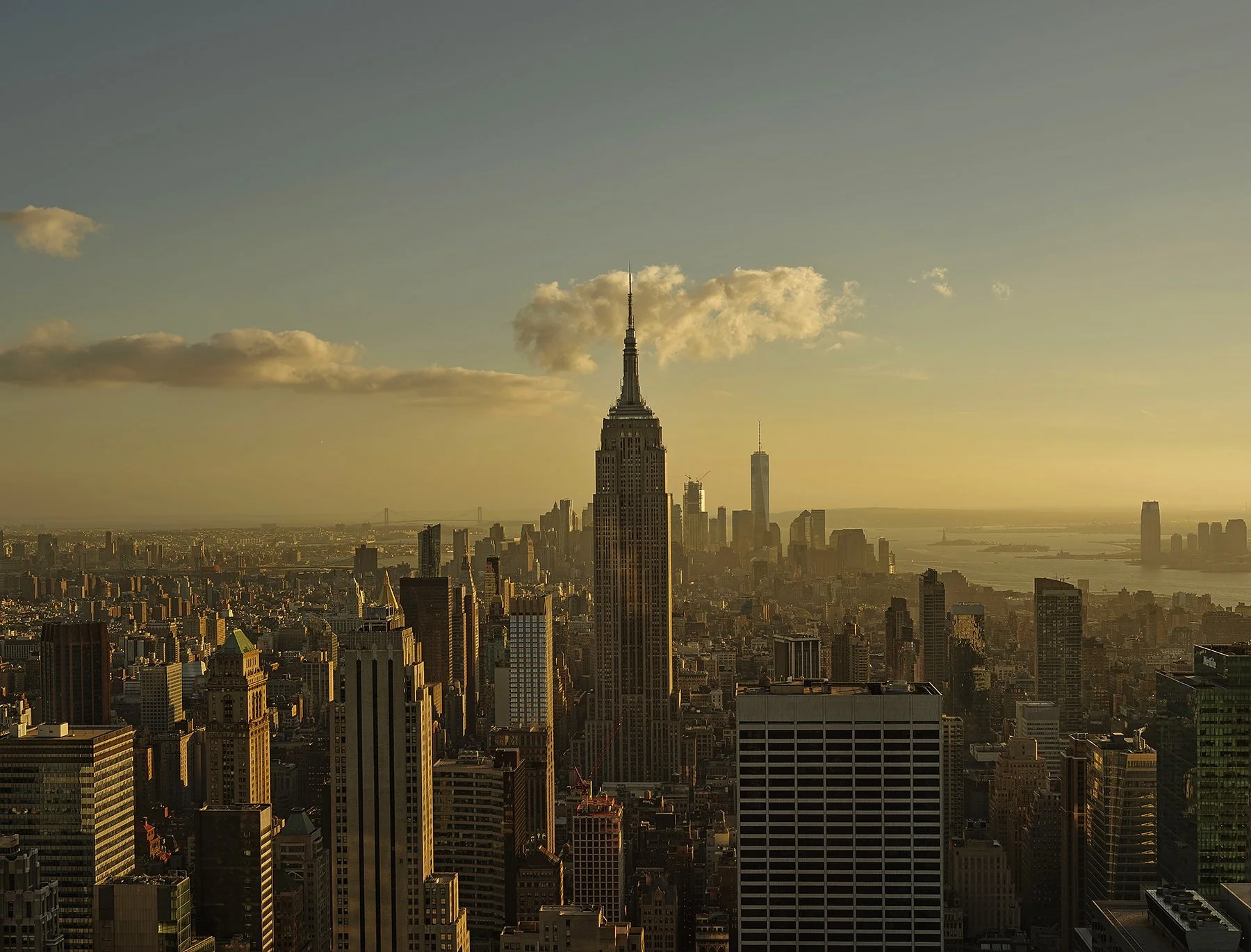 A sunset view of the New York City skyline featuring the Empire State Building and other skyscrapers, with a partly cloudy sky and the Hudson River in the background.