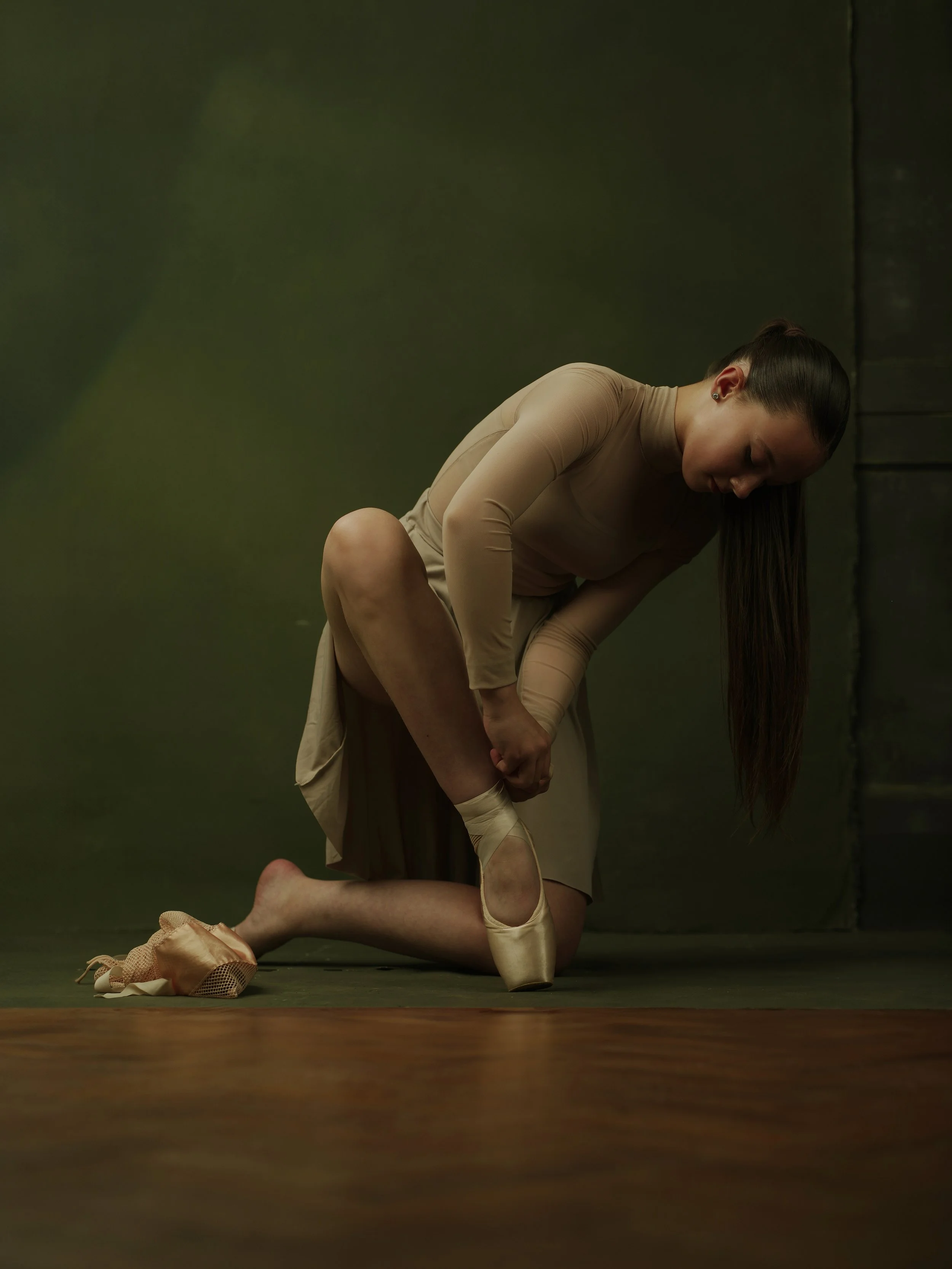 Ballet dancer adjusting her pointe shoe while kneeling on the floor in a dance studio.