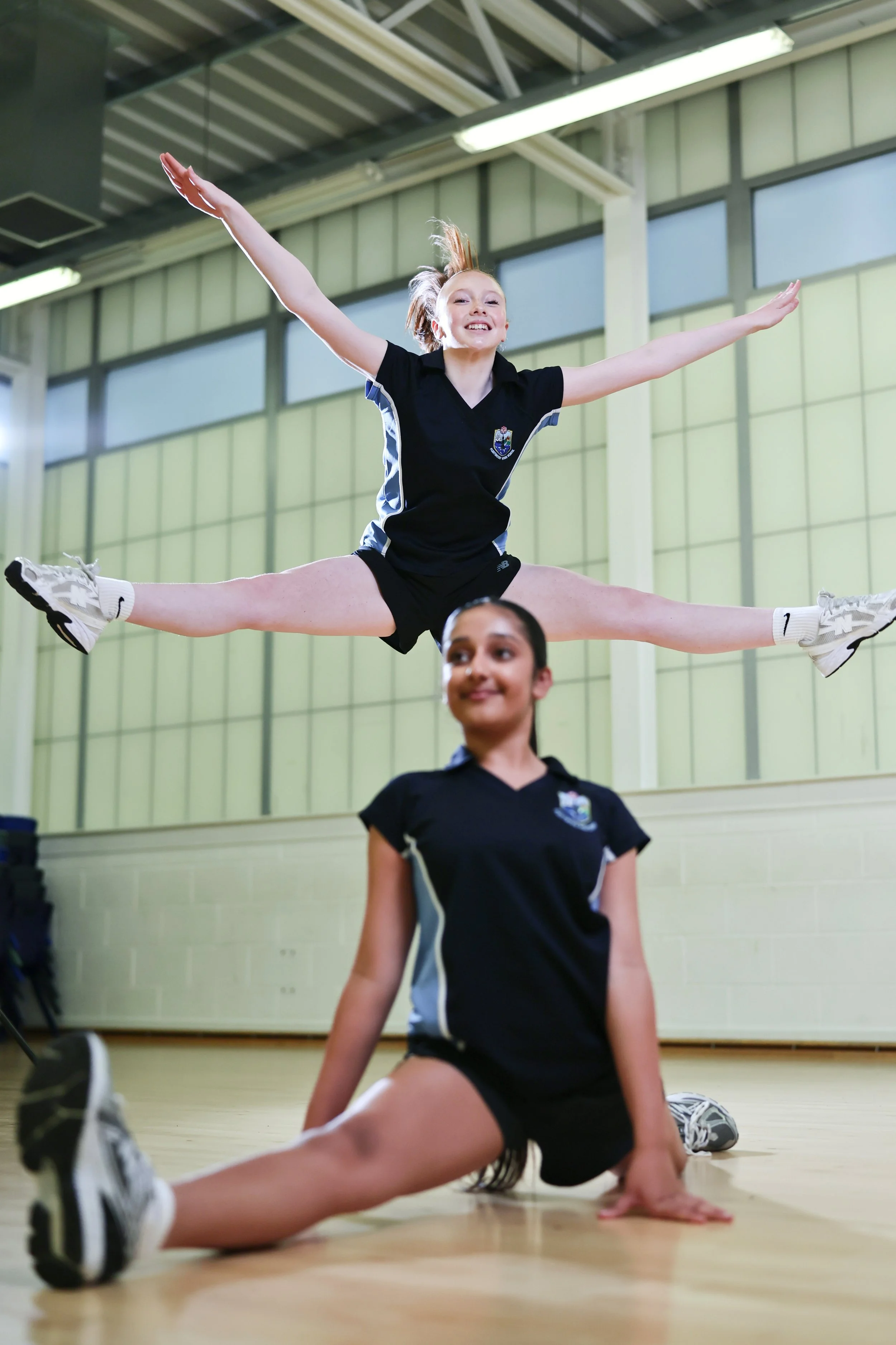 Two girls in sports uniforms practicing cheerleading or gymnastics in a gymnasium, one girl performing a jump with arms and legs extended, and the other girl kneeling or sitting on the floor in front of her.