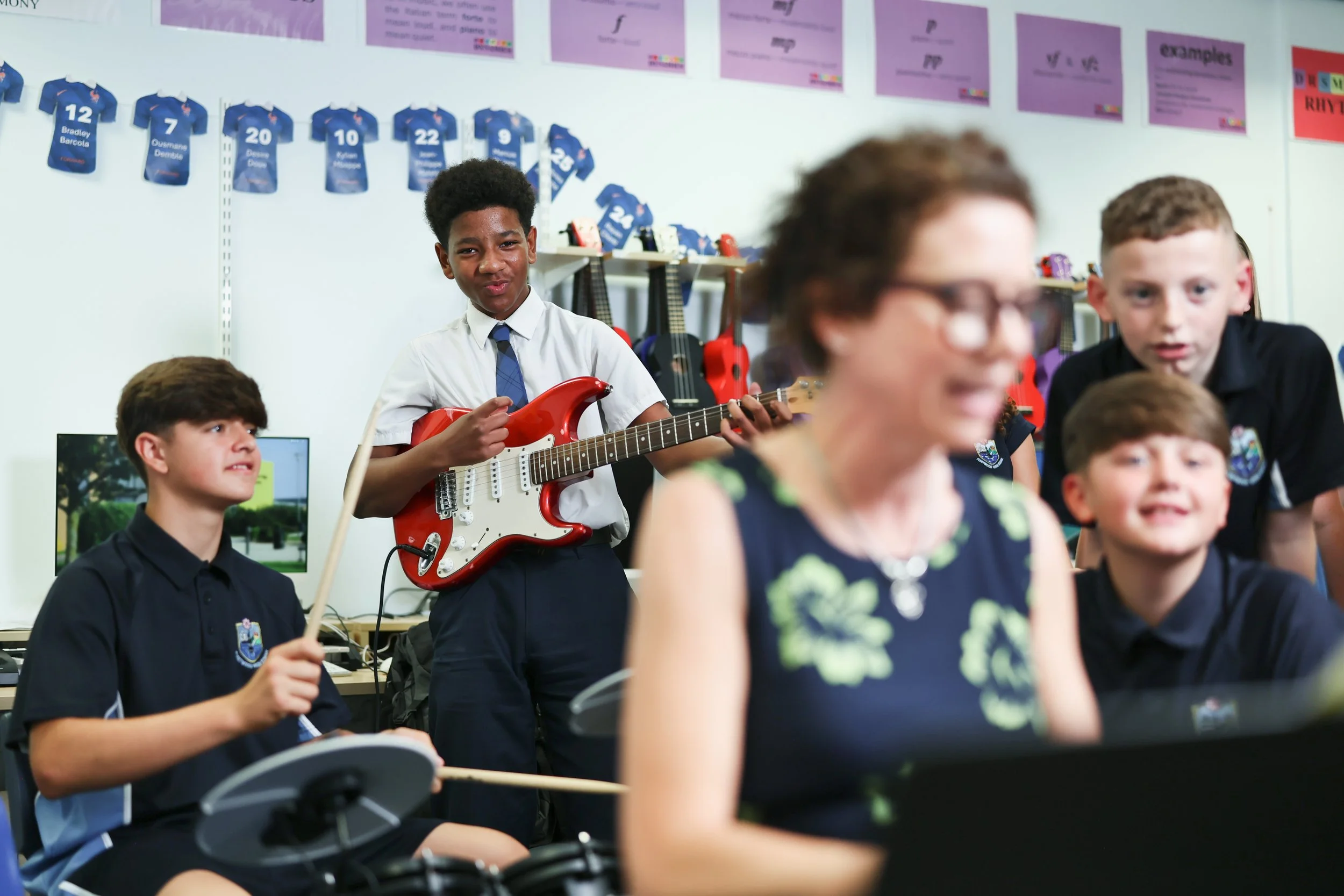 A classroom filled with students; a boy in a school uniform is playing a red electric guitar. Other students are seated, some are playing drums, and others are looking towards someone or something off-camera. The background shows posters, sports jers