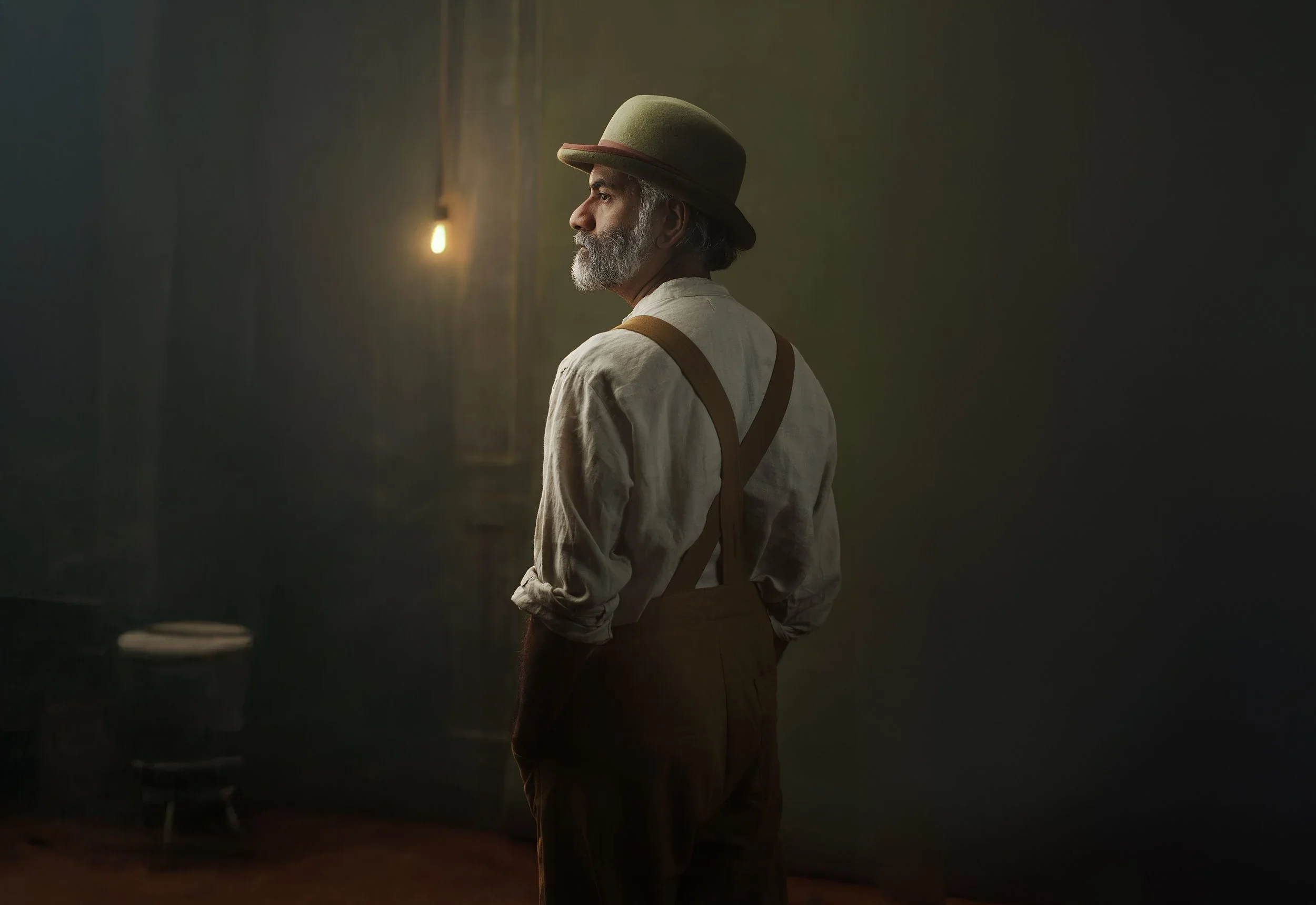 Portrait of a man in a hat and braces in front of the painted studio wall, lit by a single bulb