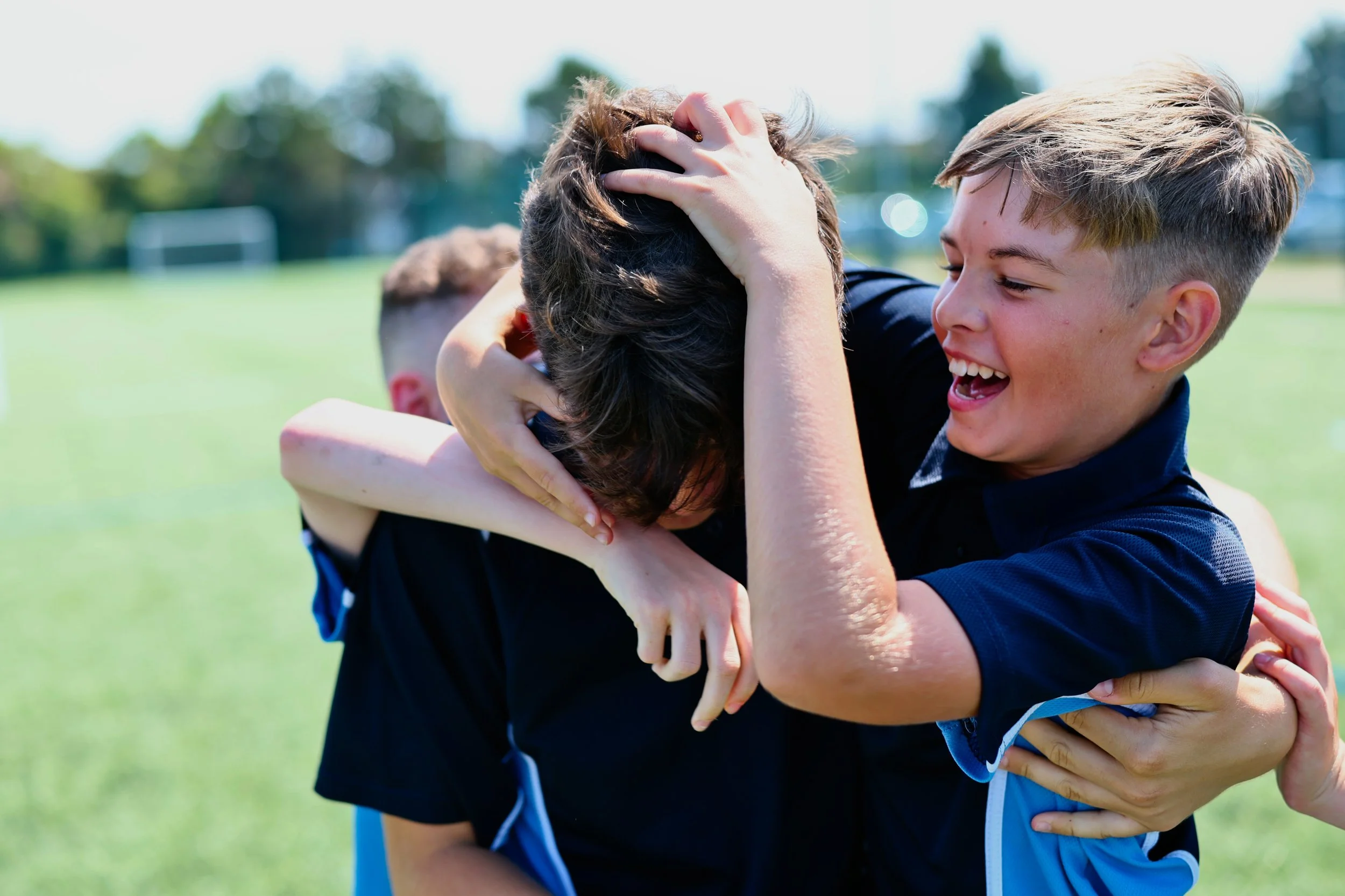 Two boys, one with blond hair and one with dark hair, are hugging passionately on a grass field on a sunny day, smiling.