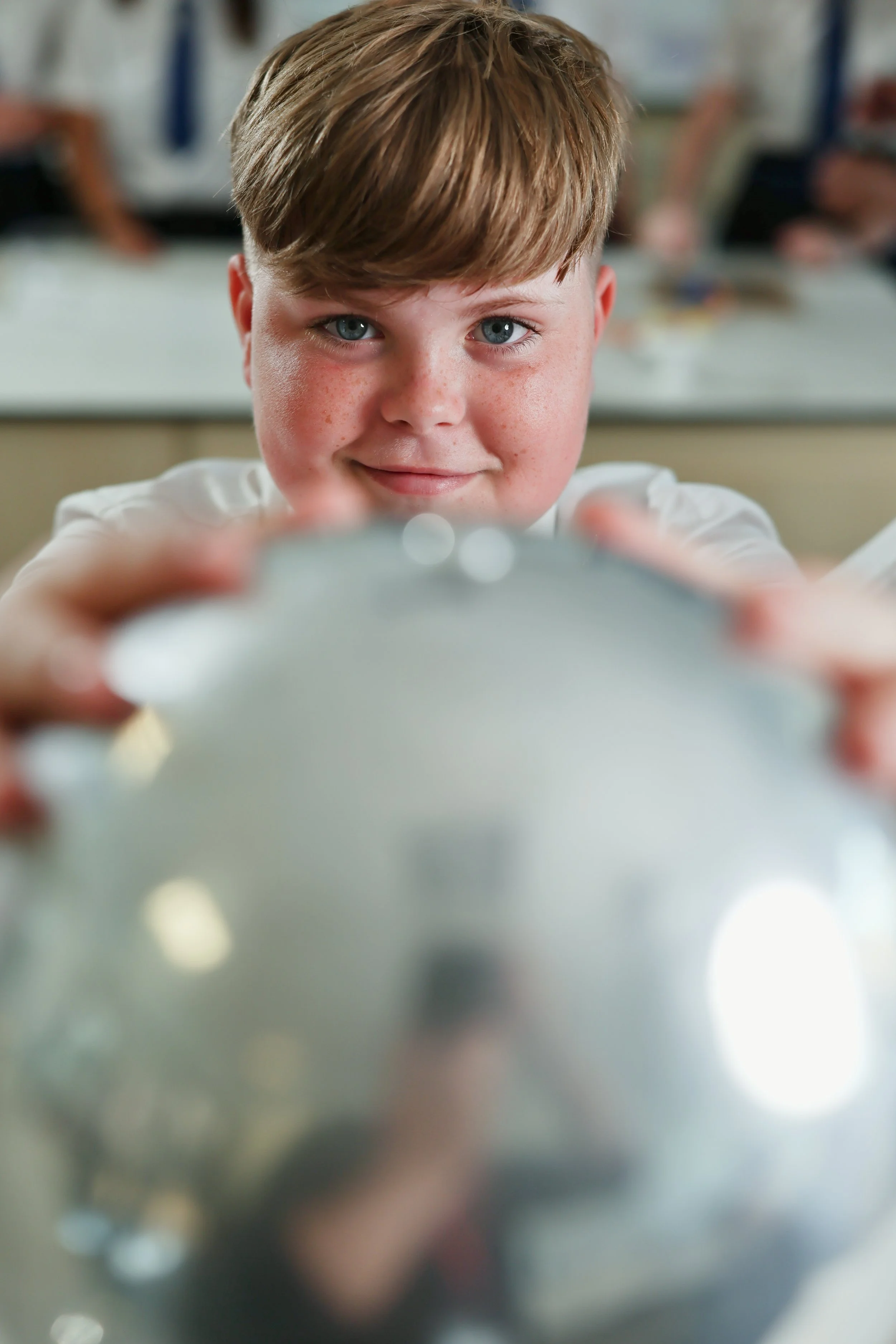 Close-up of a young boy with blonde hair and blue eyes, smiling and reaching towards the camera in a classroom setting.