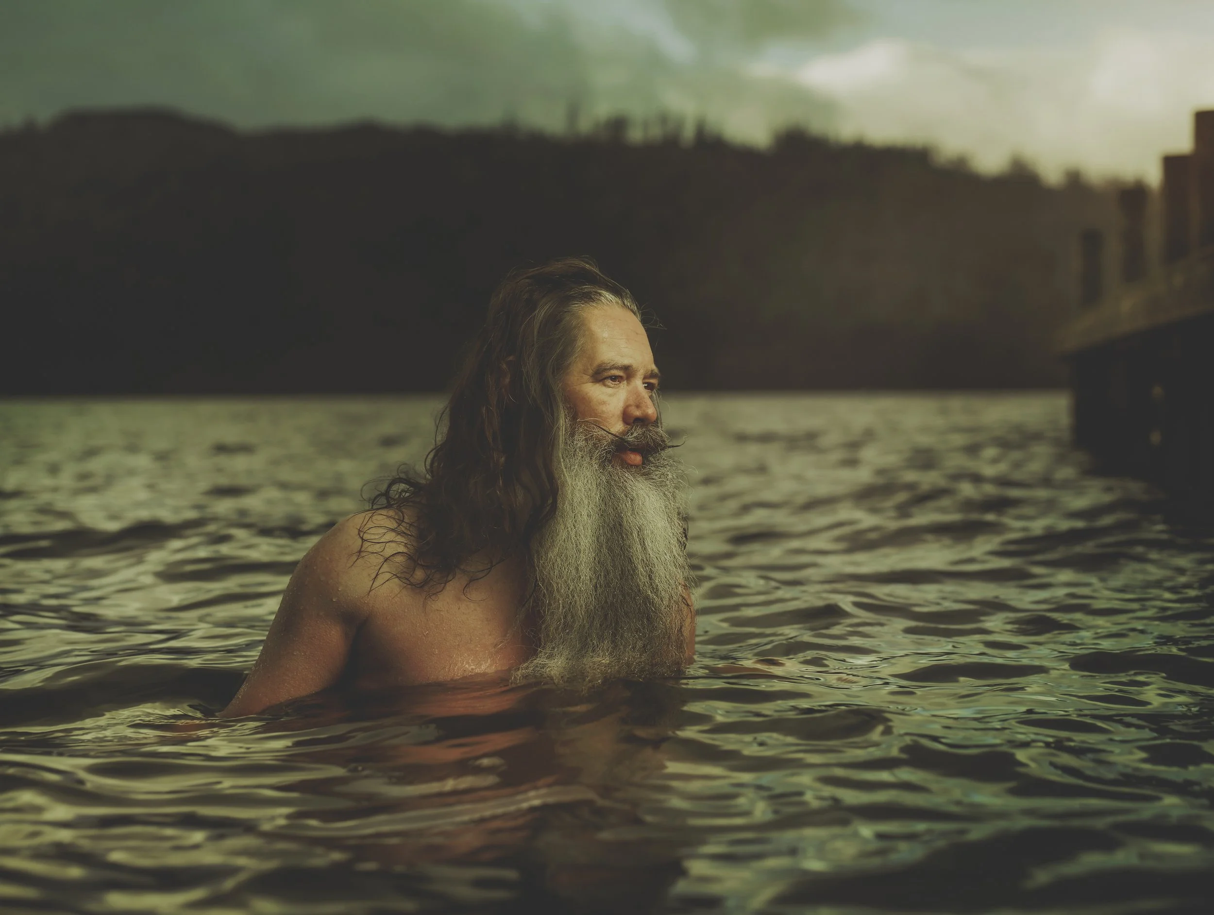 An older man with a long beard and long hair is partially submerged in water, with a background of hills and buildings under a cloudy sky.