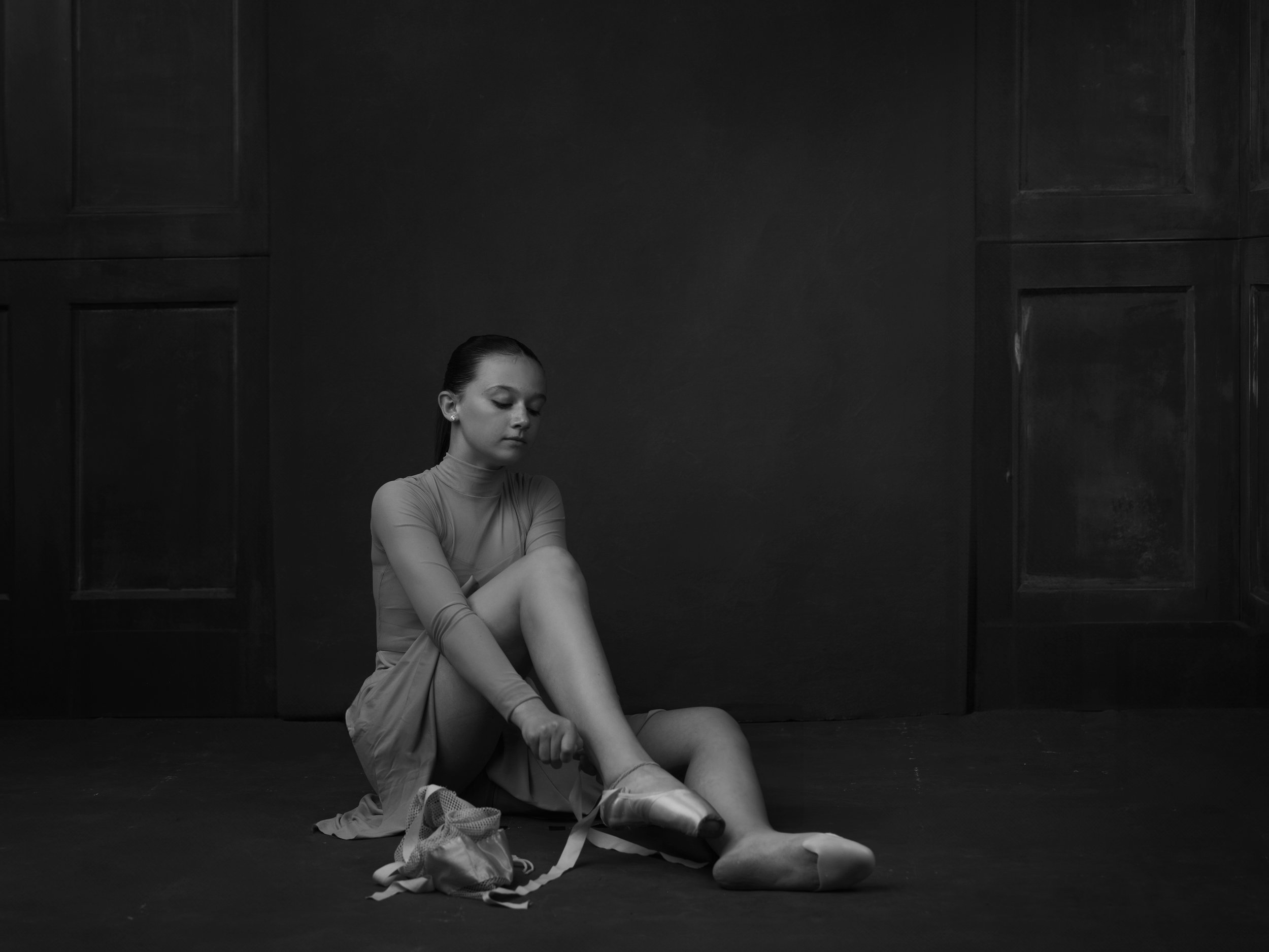 A young woman in a dance costume sits on the floor of a dark room, putting on ballet shoes.