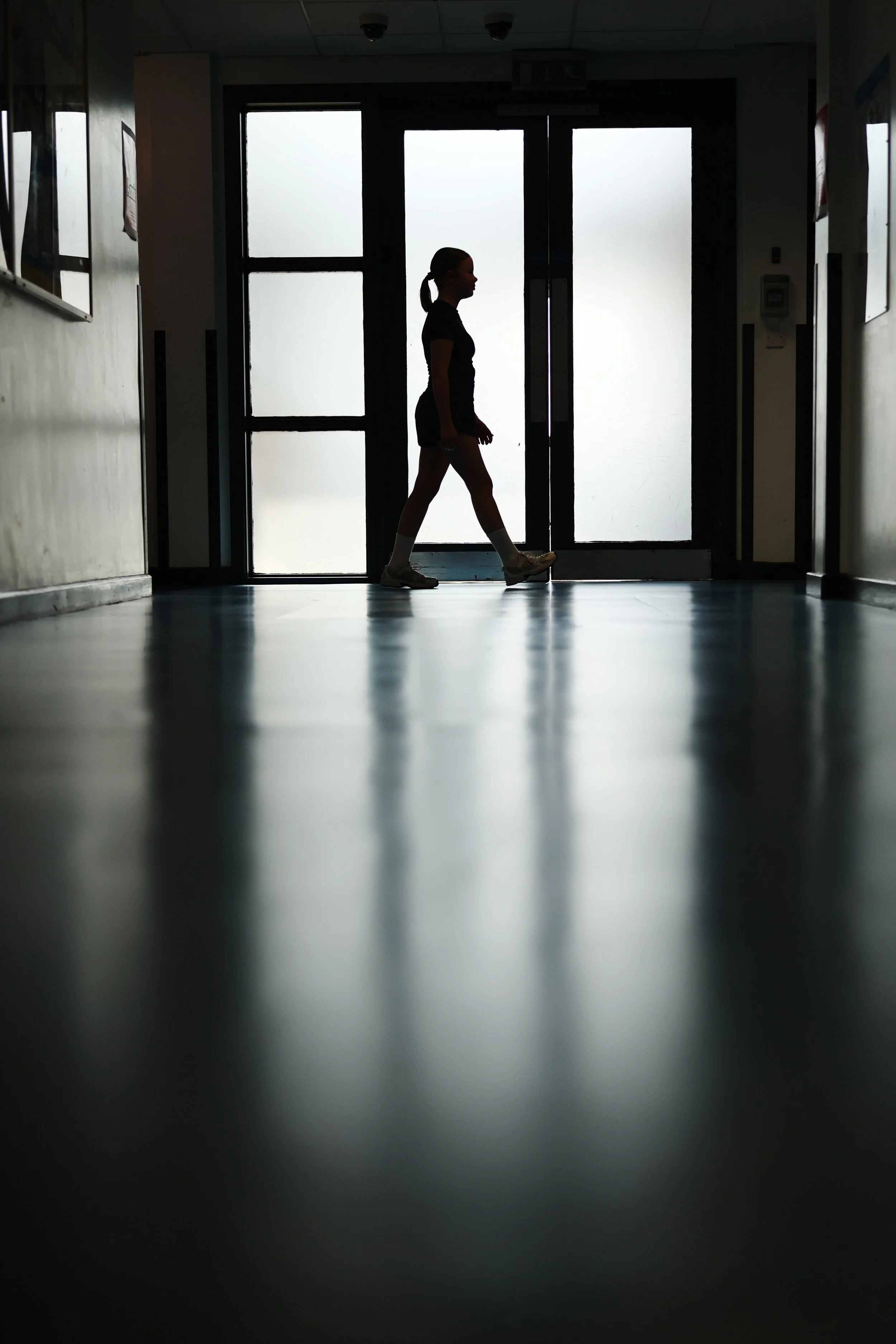 Silhouette of a girl wearing a black shirt, shorts, and sneakers walking through a hallway with frosted glass doors in the background.