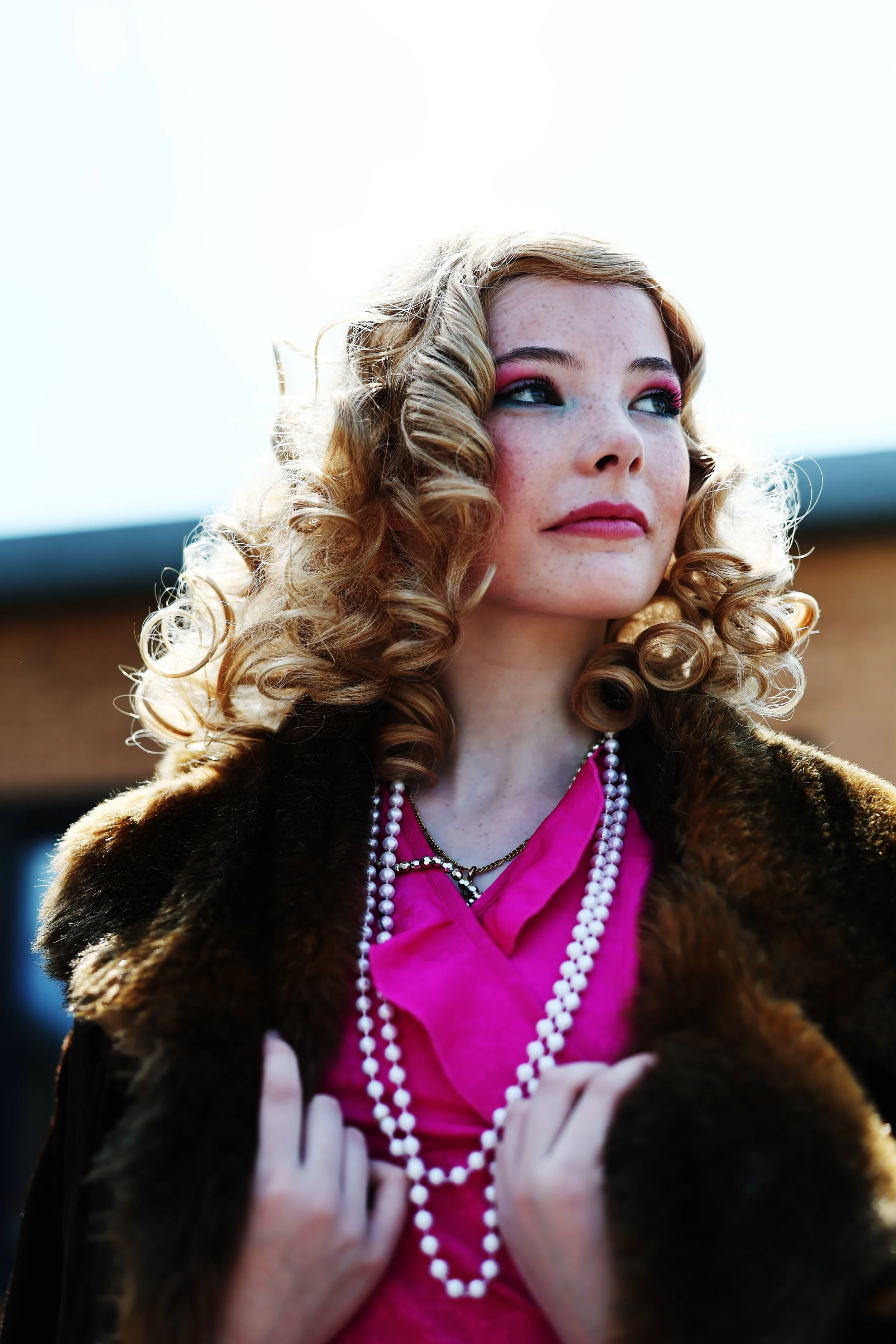 A woman with blonde, curly hair, wearing pink eyeshadow and lipstick, dressed in a bright pink top with pearl necklaces, and a fur coat, standing outdoors against a bright sky.