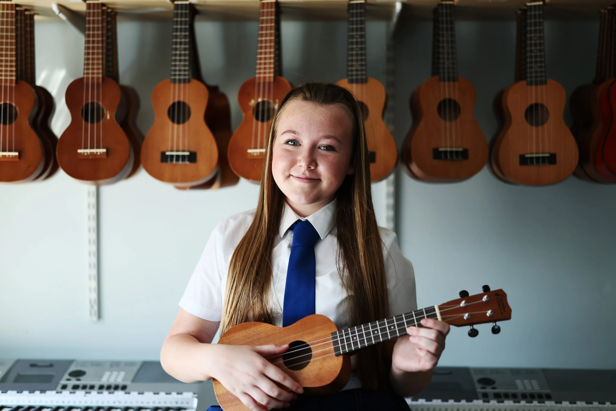 A young girl with long brown hair and a white shirt, wearing a blue tie, holding a ukulele and smiling, standing in front of a wall with hanging ukuleles.