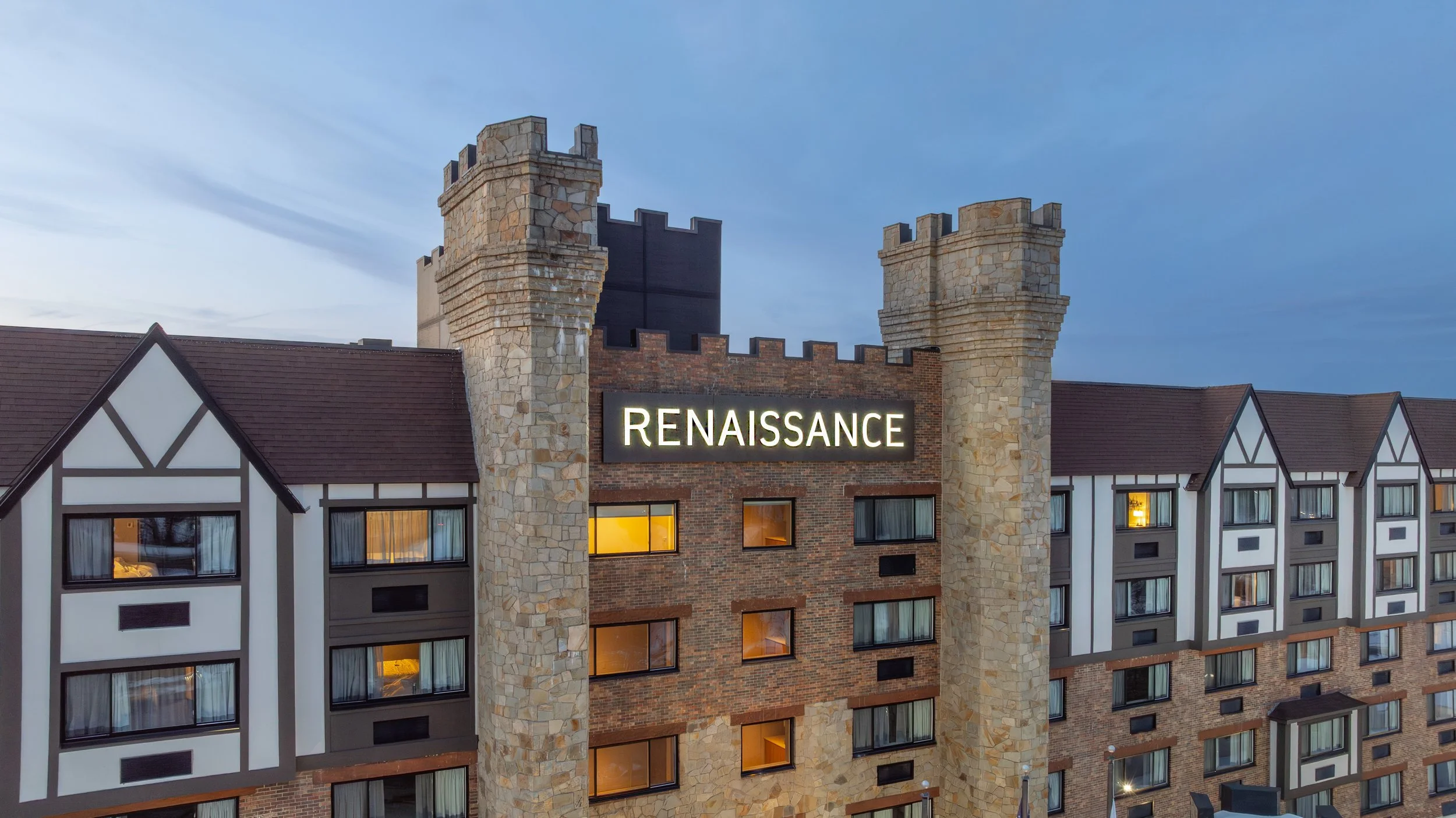 The exterior of a Renaissance hotel at dusk with illuminated windows, a large sign, and a landscaped driveway with colorful flowers and trees.