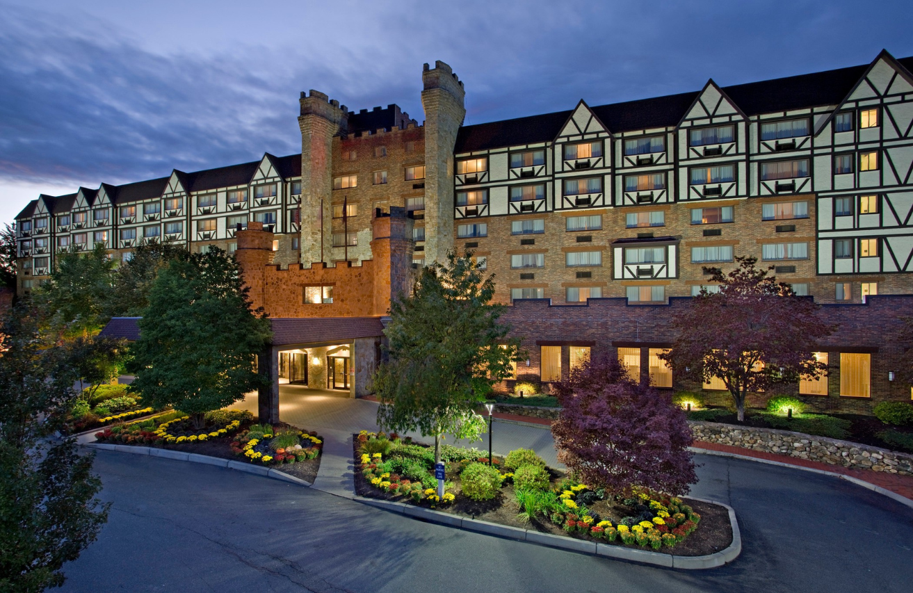 The exterior of a Sheraton hotel at dusk with illuminated windows, a large sign, and a landscaped driveway with colorful flowers and trees.