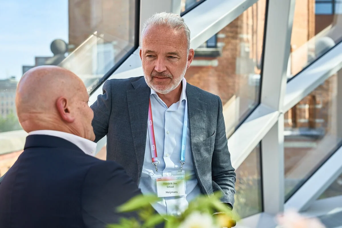 Twee mannen in gesprek, één met grijs haar en de andere zonder haren, binnen in een modern gebouw met grote ramen.