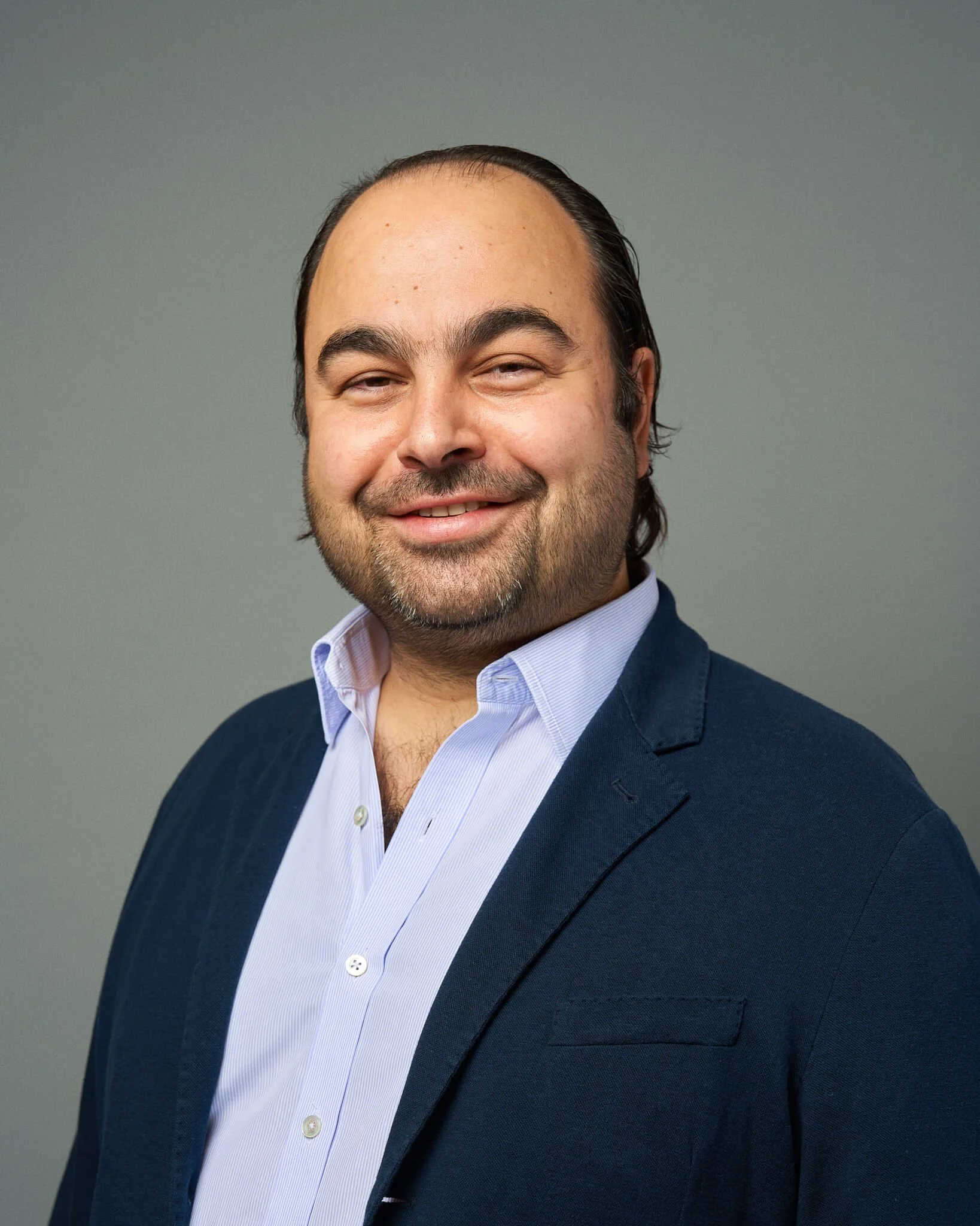 Portret van een man met een brede glimlach, donker haar en een volle baard, gekleed in een lichtblauwe overhemd en een donkerblauwe blazer, tegen een effen grijze achtergrond.