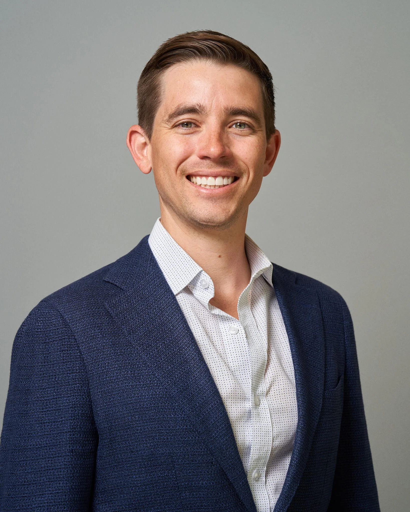 Portret van een lachende man met bruin haar in een net pak, tegen een effen achtergrond.