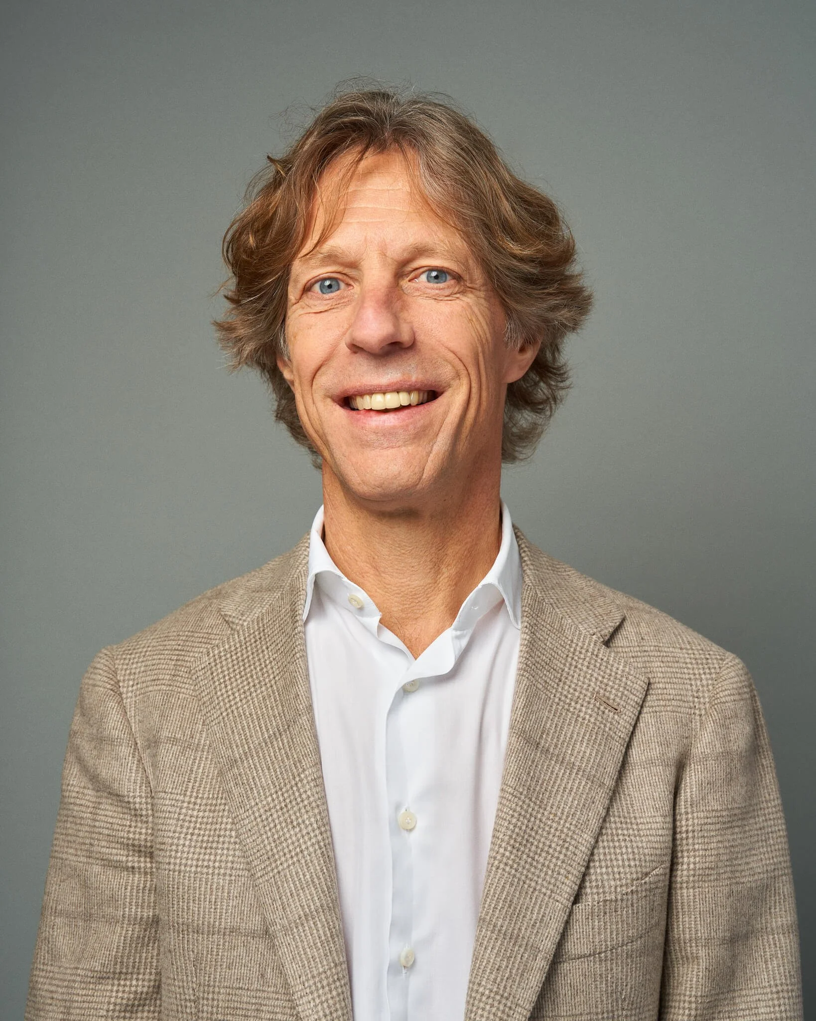 Portret van een glimlachende man met bruin, golvend haar, gekleed in een witte overhemd en een beige geruite jas, voor een grijze achtergrond.