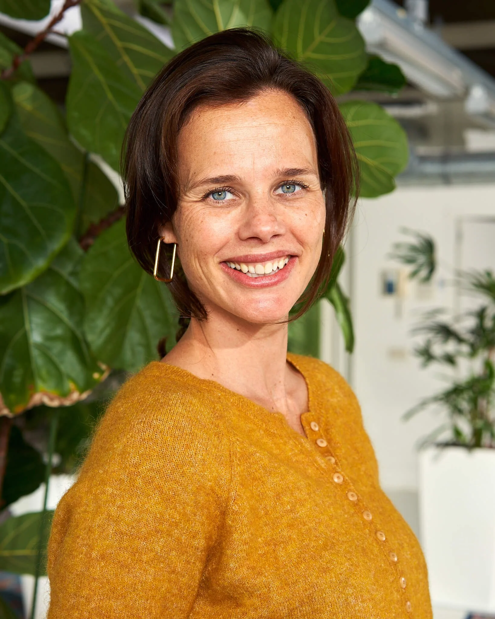 Portret van een lachende vrouw met kort bruin haar, blauwe ogen, grote oorbellen, draagt een gele trui, op de achtergrond grote groene bladeren en een interieur met planten. Gefotografeerd door een professionele portretfotograaf.