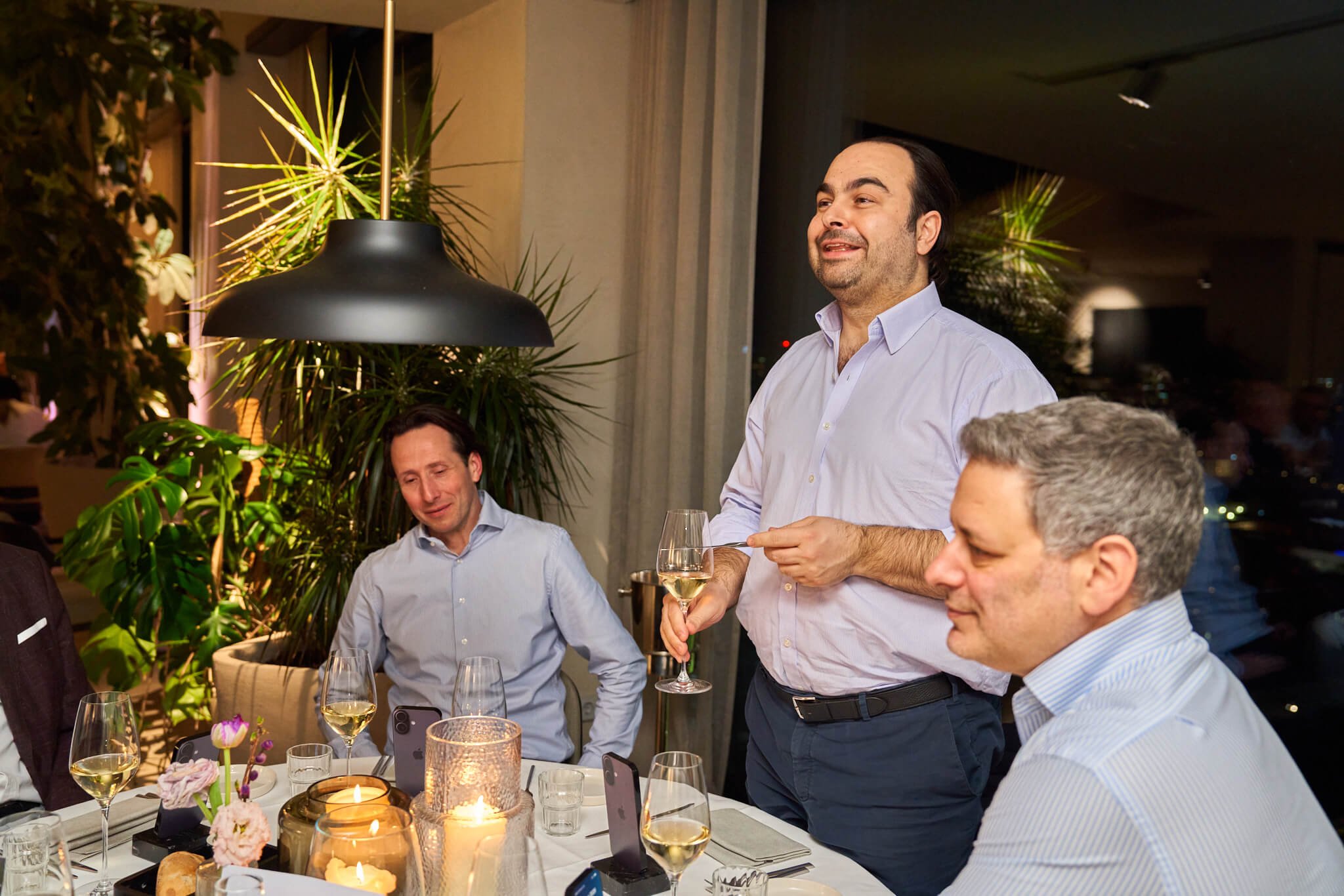 Een man staat en spreekt tijdens een diner, hij houdt een glas witte wijn vast. Andere mensen zitten aan de tafel, die is versierd met kaarsen, bloemen en bestek. Het diner vindt plaats in een sfeervolle, moderne ambiance met grote planten enhelder v