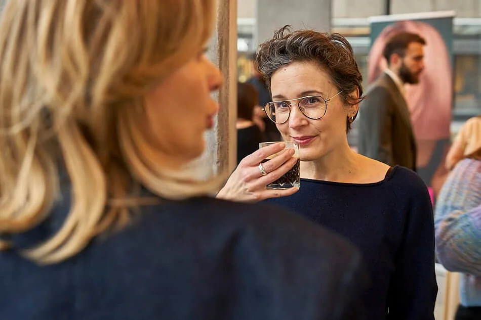 Twee vrouwen die met elkaar praten en drinken op een evenement, op de achtergrond is een man met een baard en een poster zichtbaar.