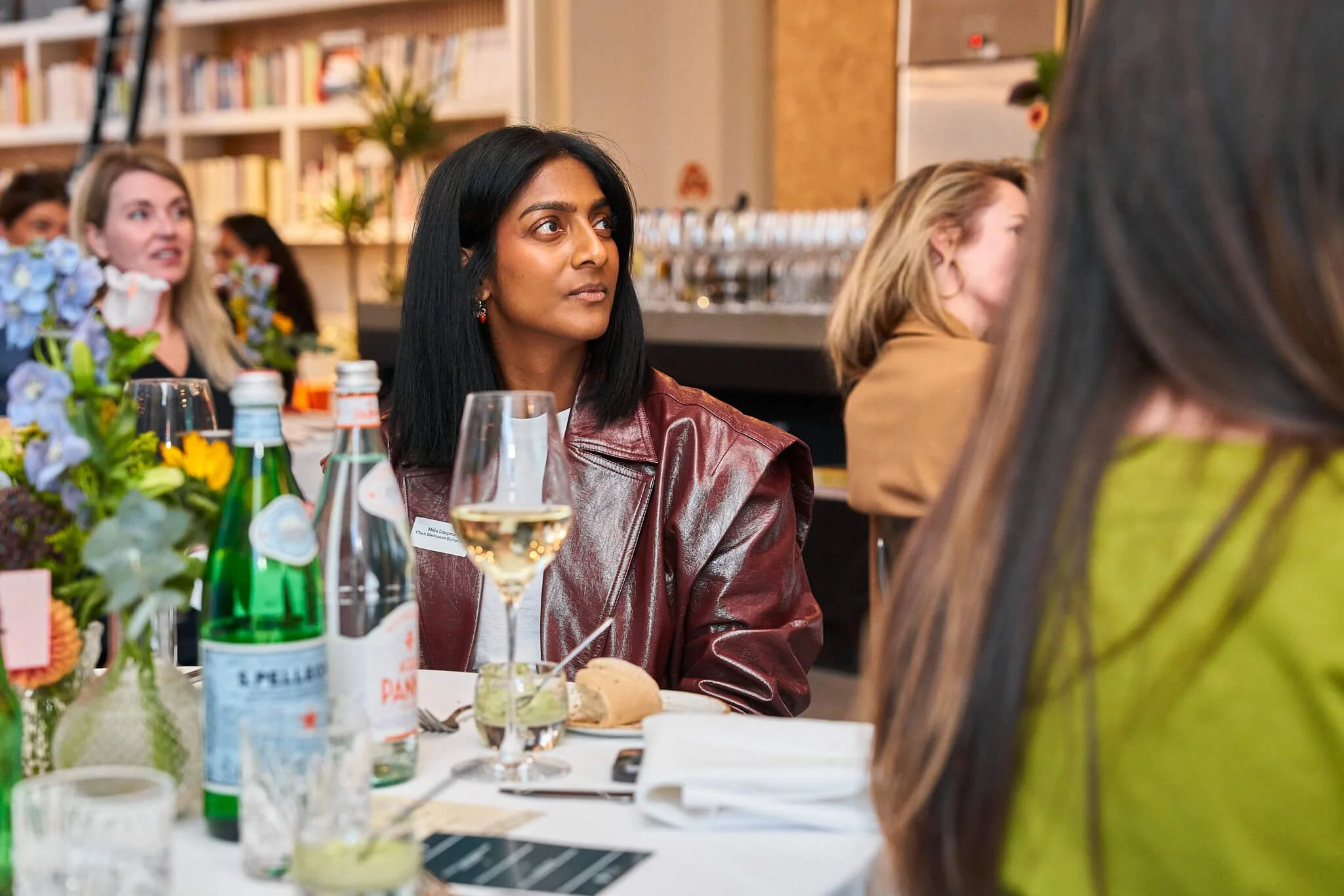 Een vrouw met zwarte haar, gekleed in een rode leren jas, zit aan een tafel tijdens een bijeenkomst of diner, met bloemstukken en drankjes op tafel.