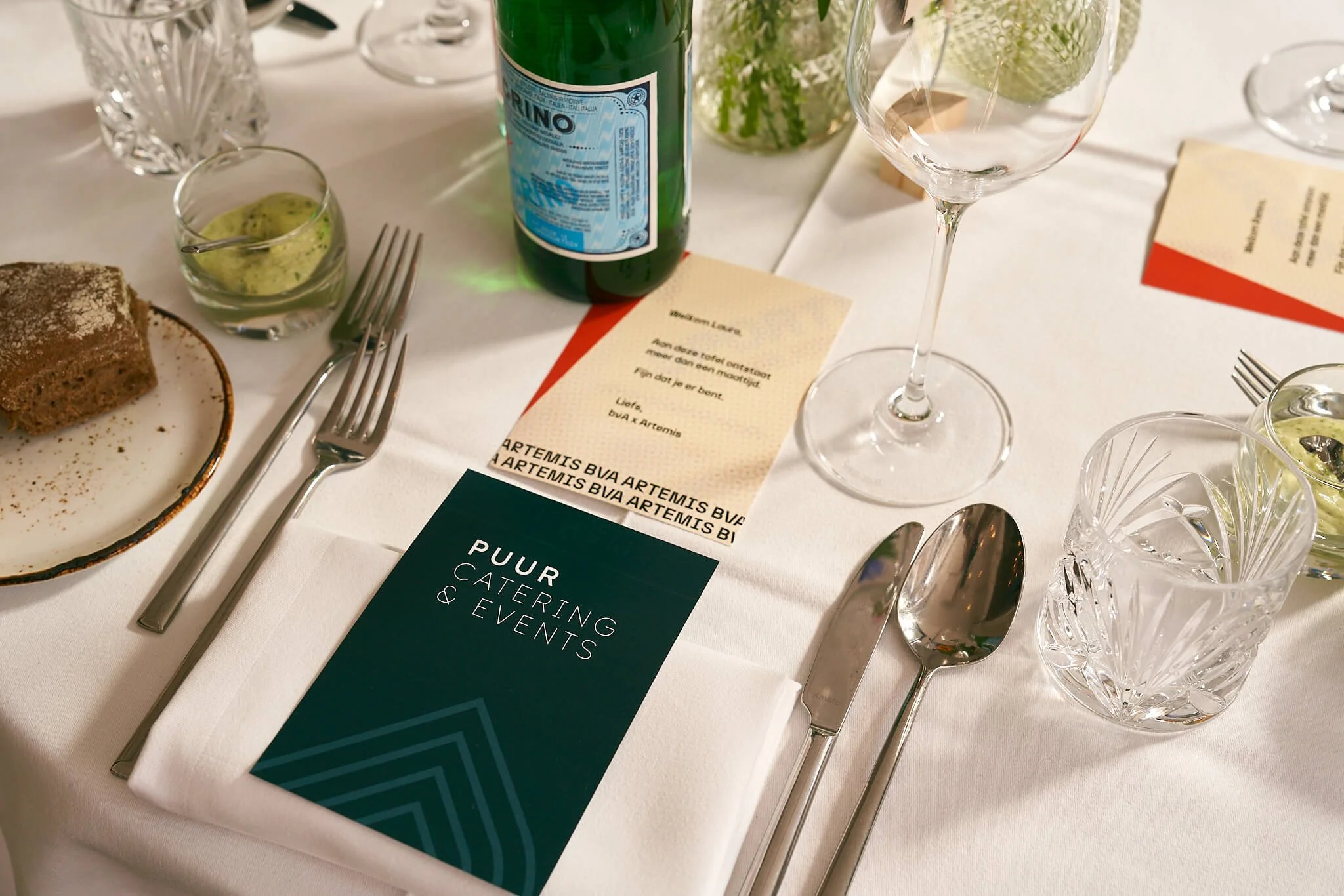 Een gedekte tafel met glazen, bestek, een stuk cake, een fles water, en een menu voor een catering- en evenementenbureau, versierd met bloemen.