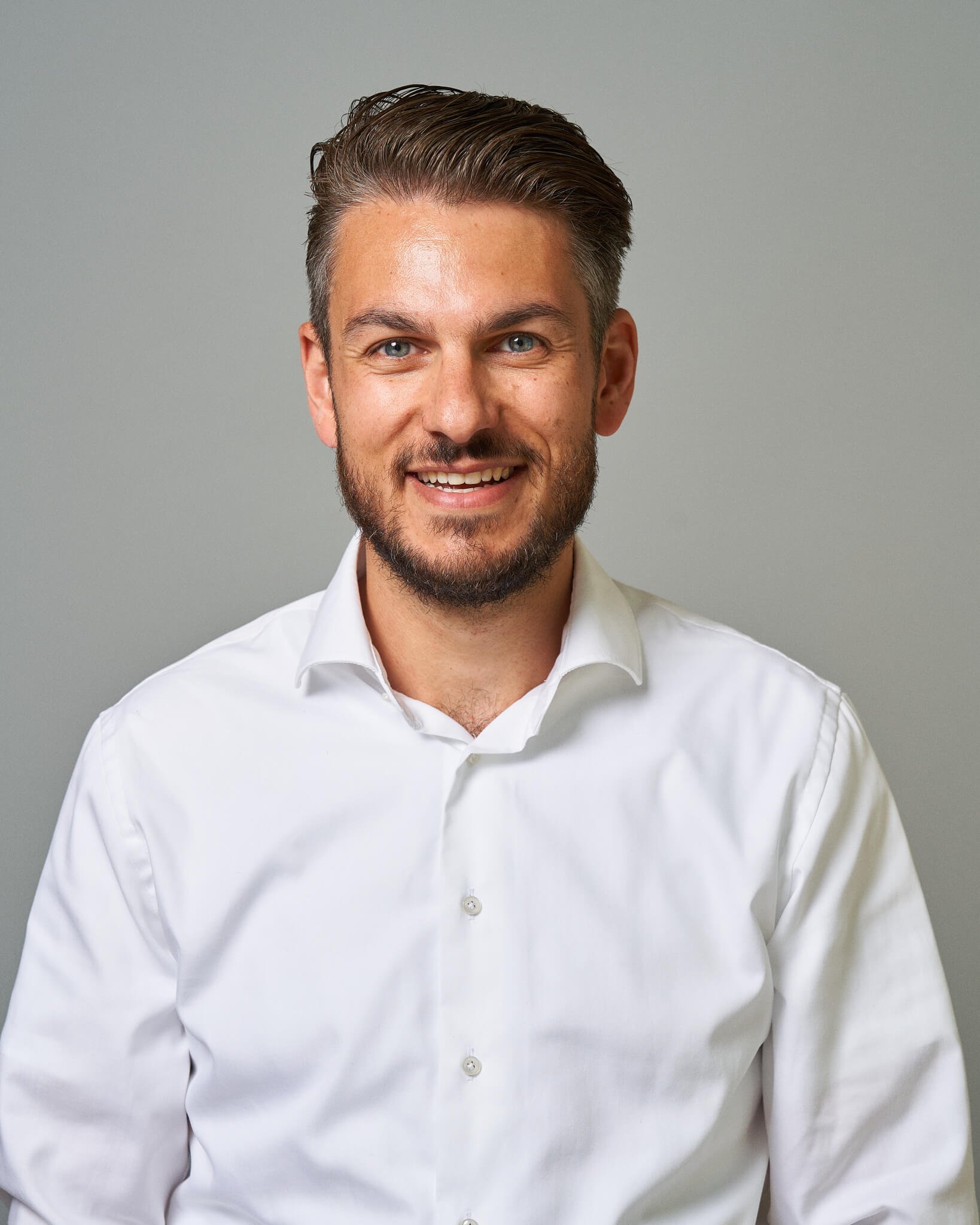 Portret van een glimlachende man met donker haar, een baard en blauwe ogen, gekleed in een witte blouse op een neutrale achtergrond.
