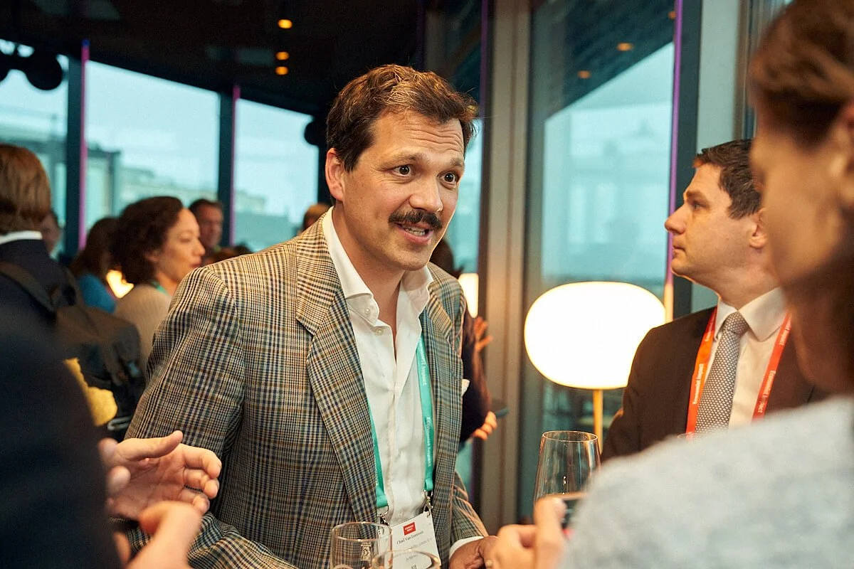 Man in een lichtbruin geruit jasje met witte blouse die spreekt tijdens een netwerkbijeenkomst, omringd door anderen die luisteren, met wijn glazen in de hand.