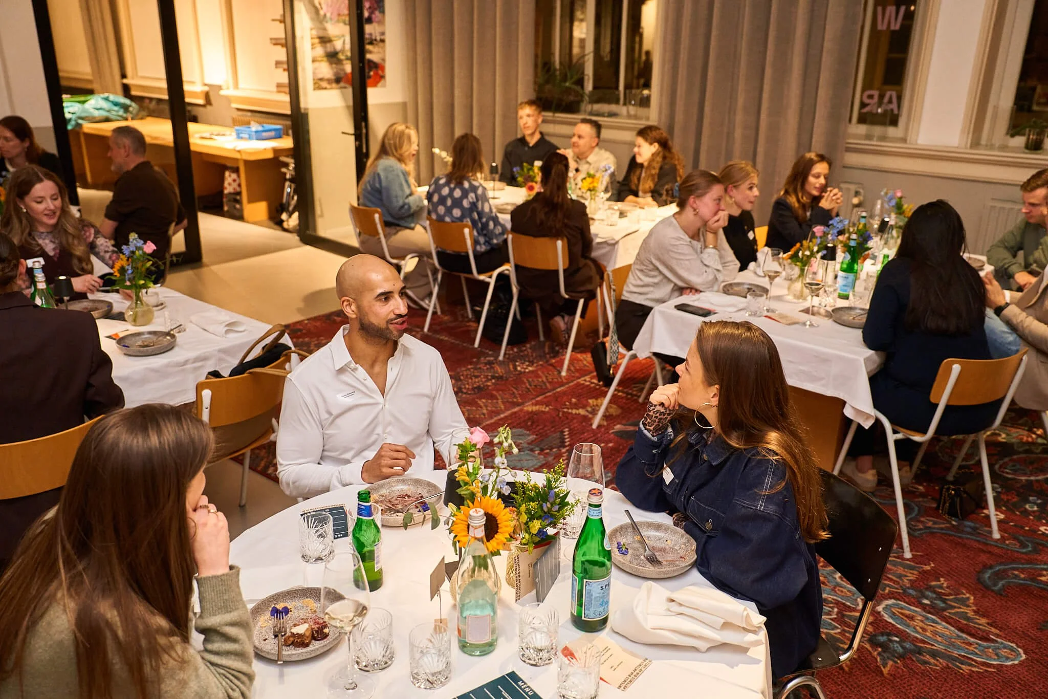Een groep mensen zit aan tafels tijdens een diner of evenement in een gezellig, goed verlichte ruimte.