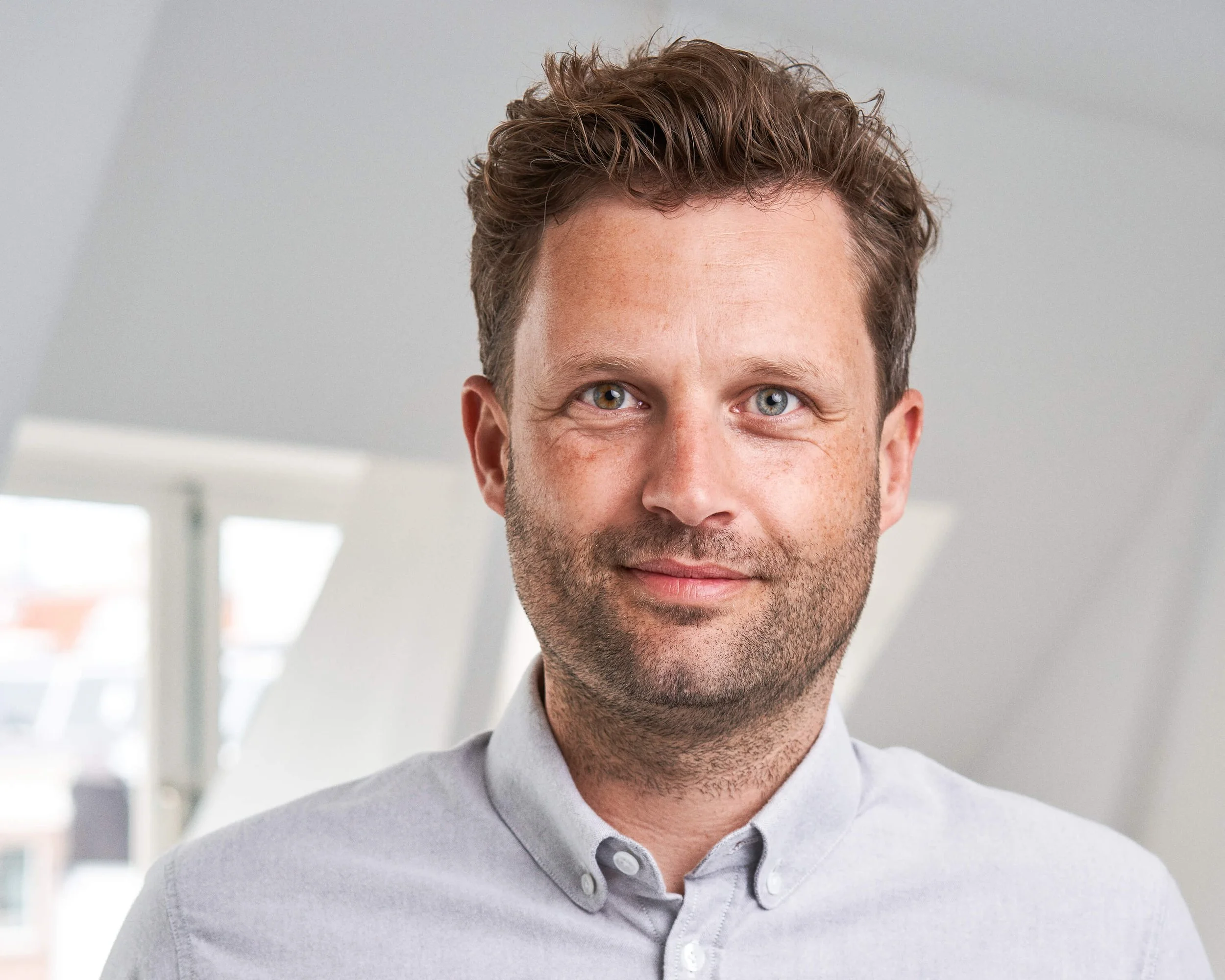 Portret van een man met lichte huid, bruin haar en blauwe ogen, die glimlacht, gekleed in een lichtgrijs overhemd, binnen in een lichte kamer met grote ramen.