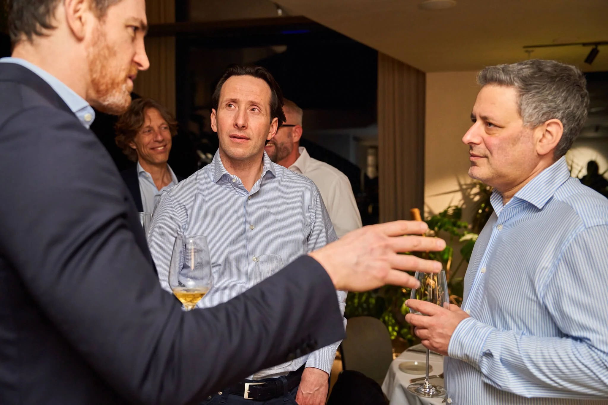 Vijf mannen in een restaurant of feestzaal die in gesprek zijn, met drie prominente en twee op de achtergrond.