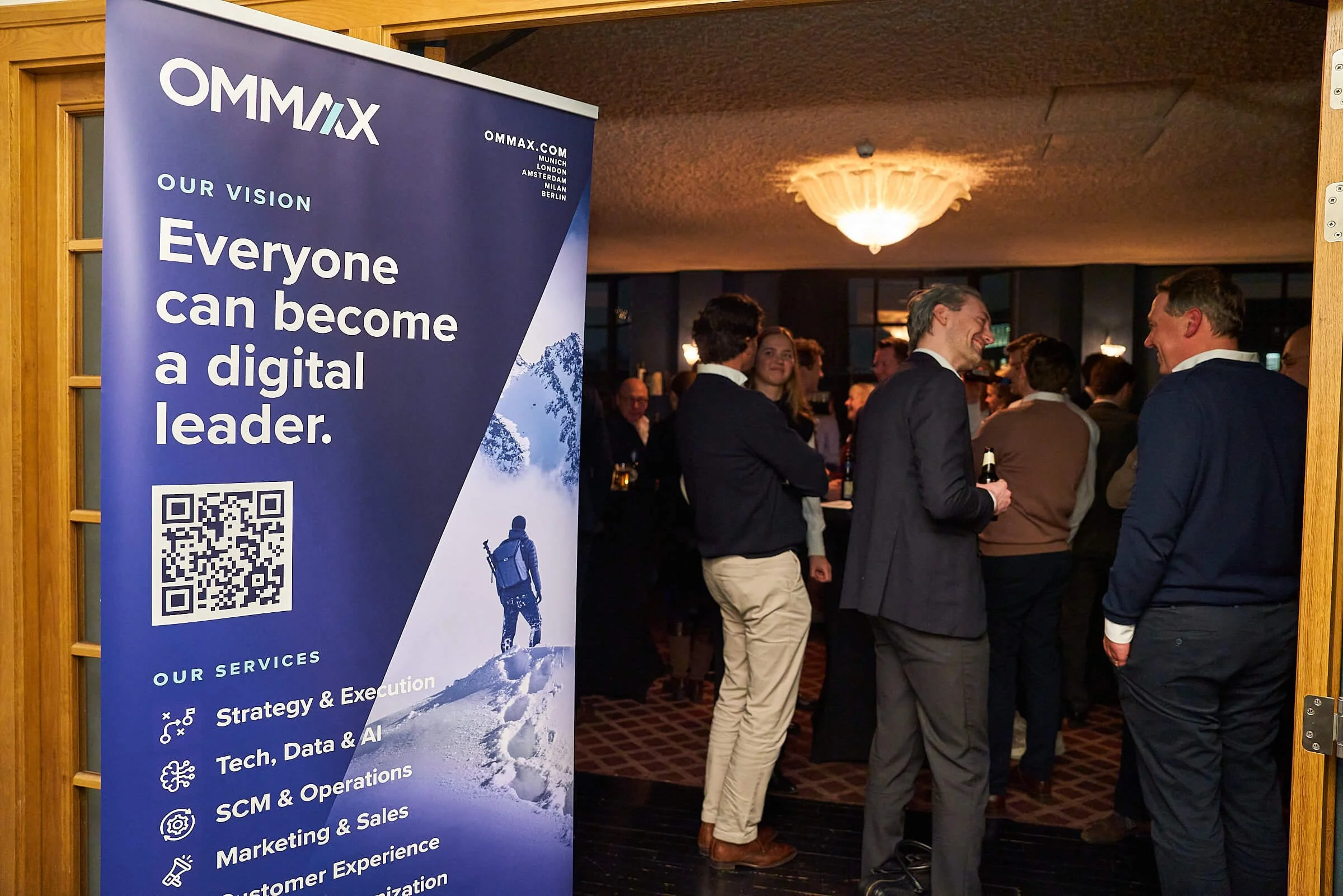 Een groep mensen in conversatie in een binnenruimte met een plafondlamp, naast een grote blauwe banner met de tekst 'Everyone can become a digital leader' en een QR-code, bij een evenement of bijeenkomst.